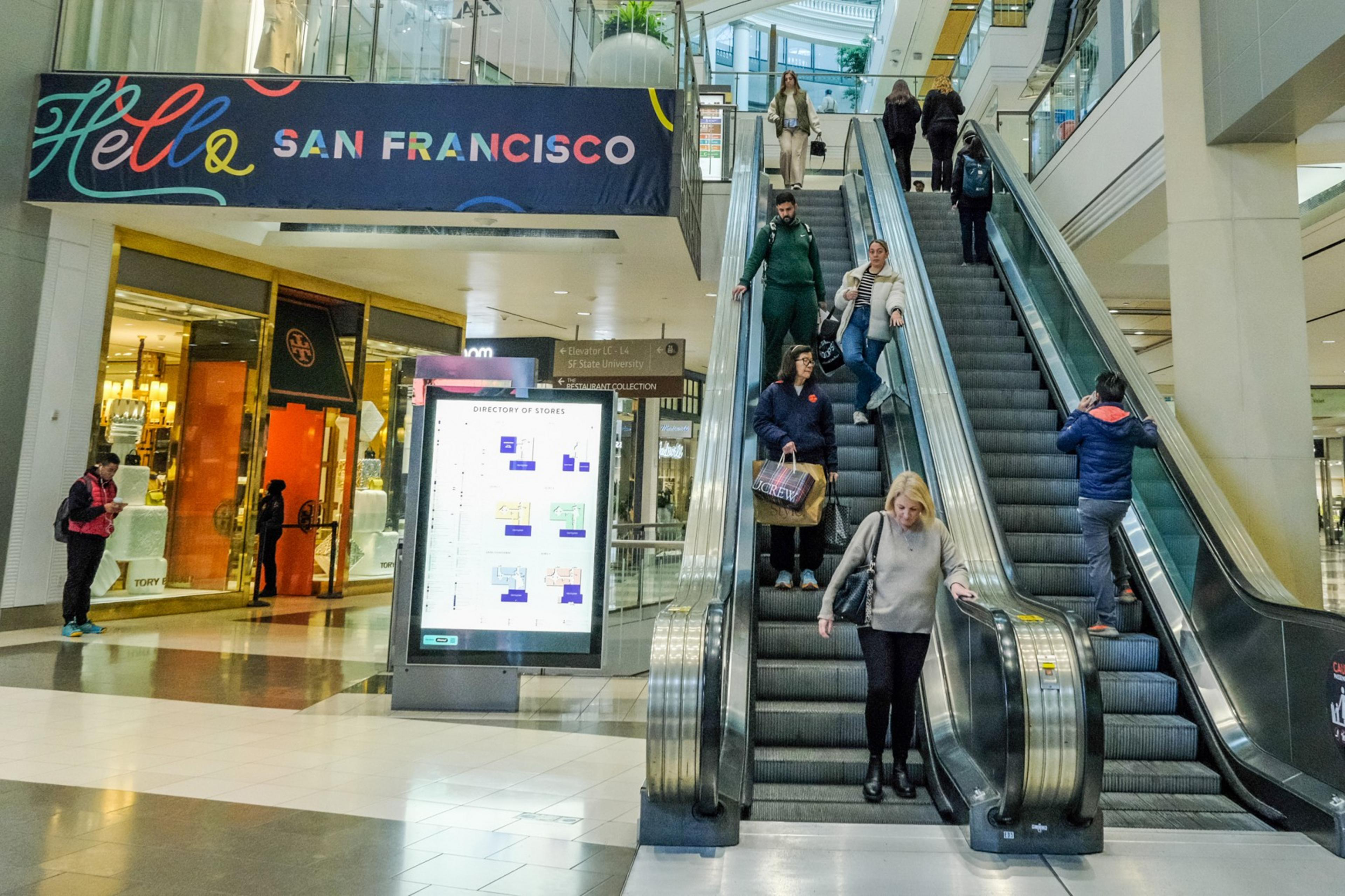 People are using an escalator in a modern indoor shopping mall with stores and a digital directory.