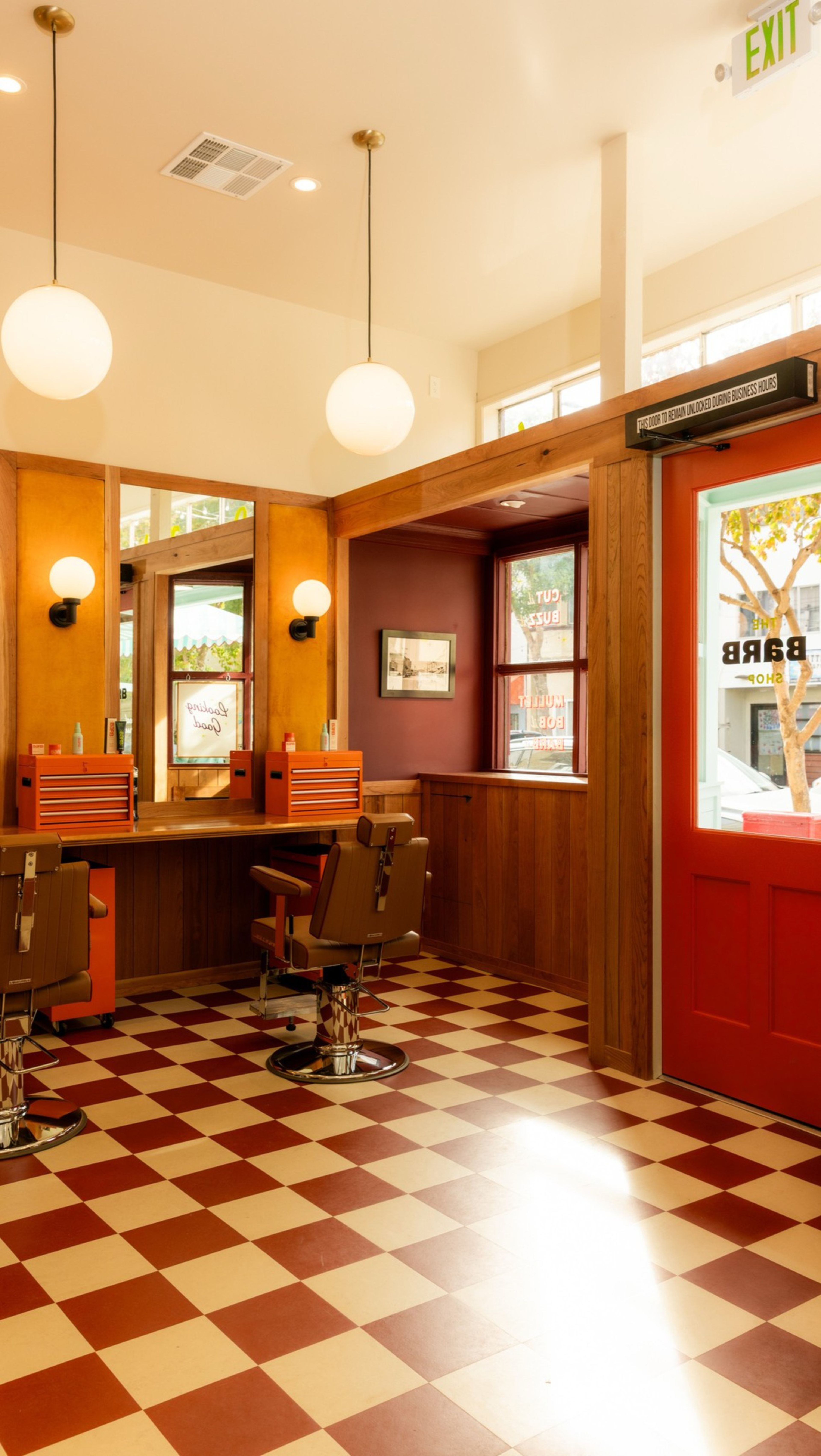 A barber shop interior with checkered red and cream floor tiles, two brown barber chairs, wooden counters, round wall lamps, and a bright red door.