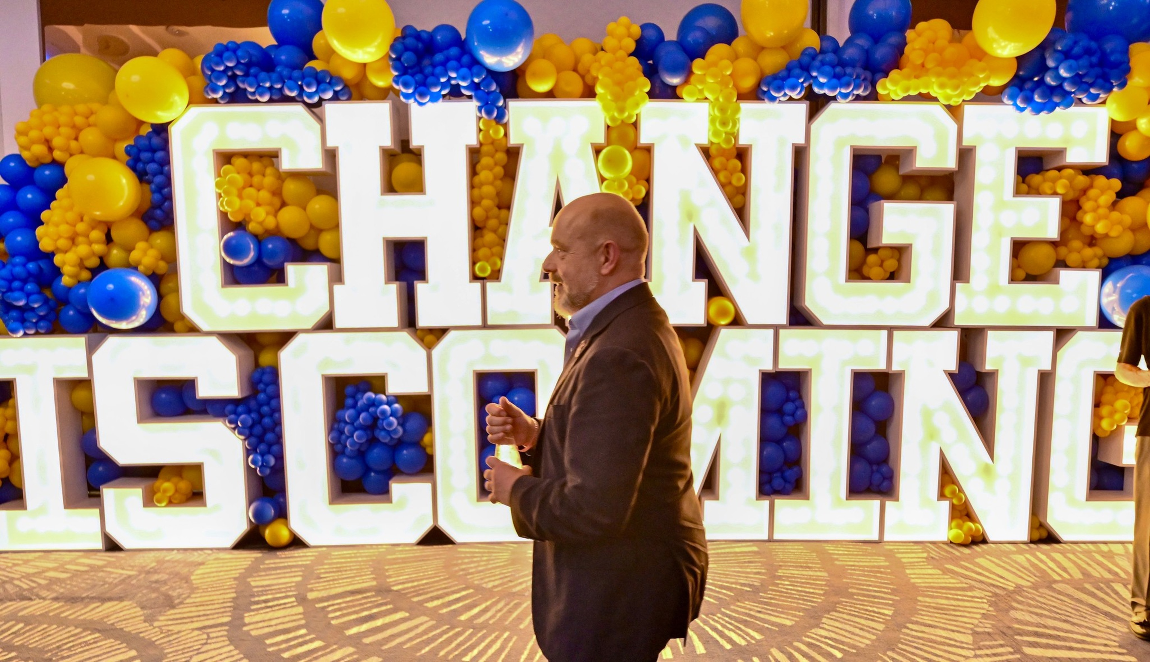A man in a suit walks past large illuminated letters reading “CHANGE IS COMING,” surrounded by blue and yellow balloons.