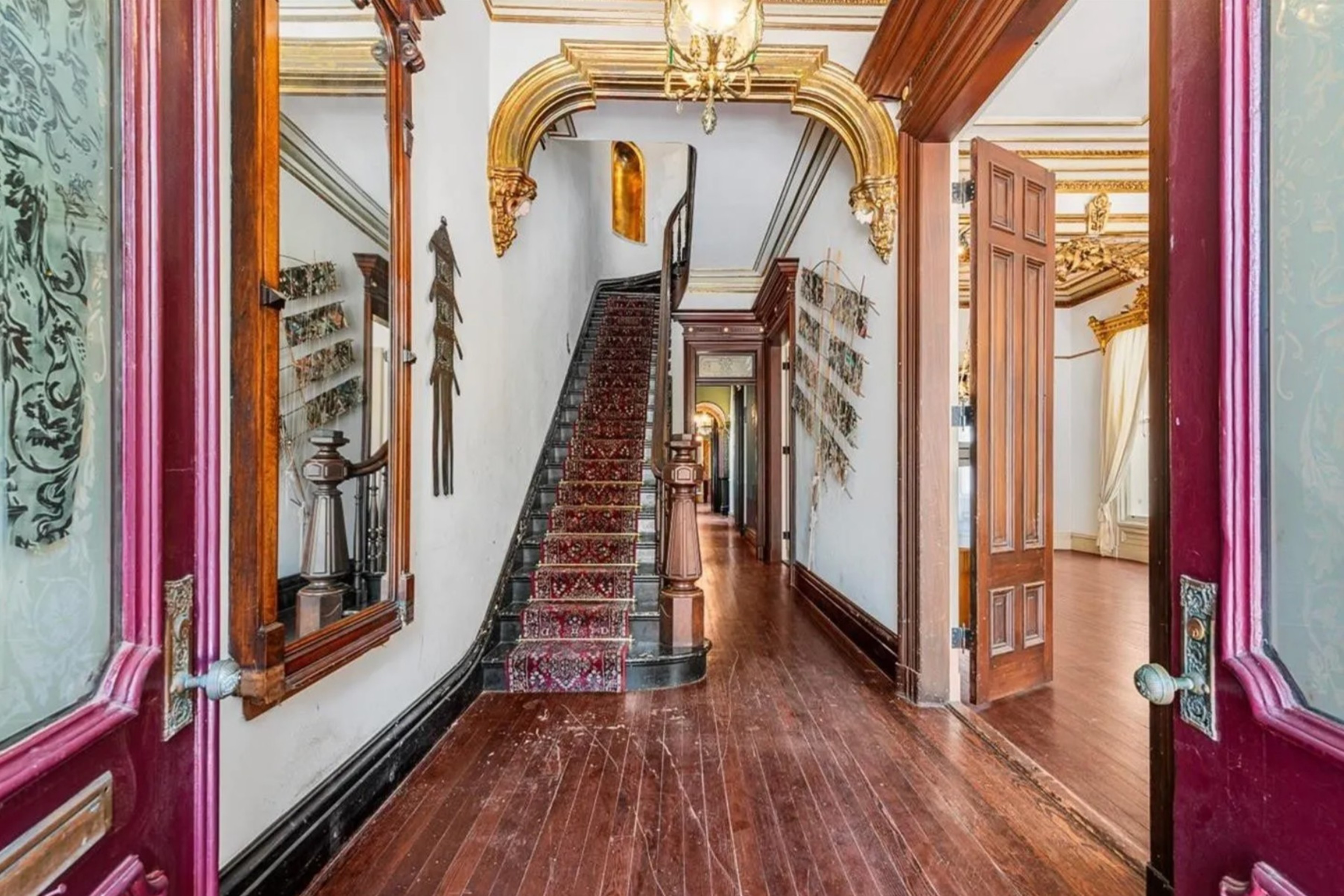 The image shows an ornate, vintage hallway with wooden floors, a carpeted staircase, elaborate gold-trimmed ceilings, and richly detailed door frames and moldings.