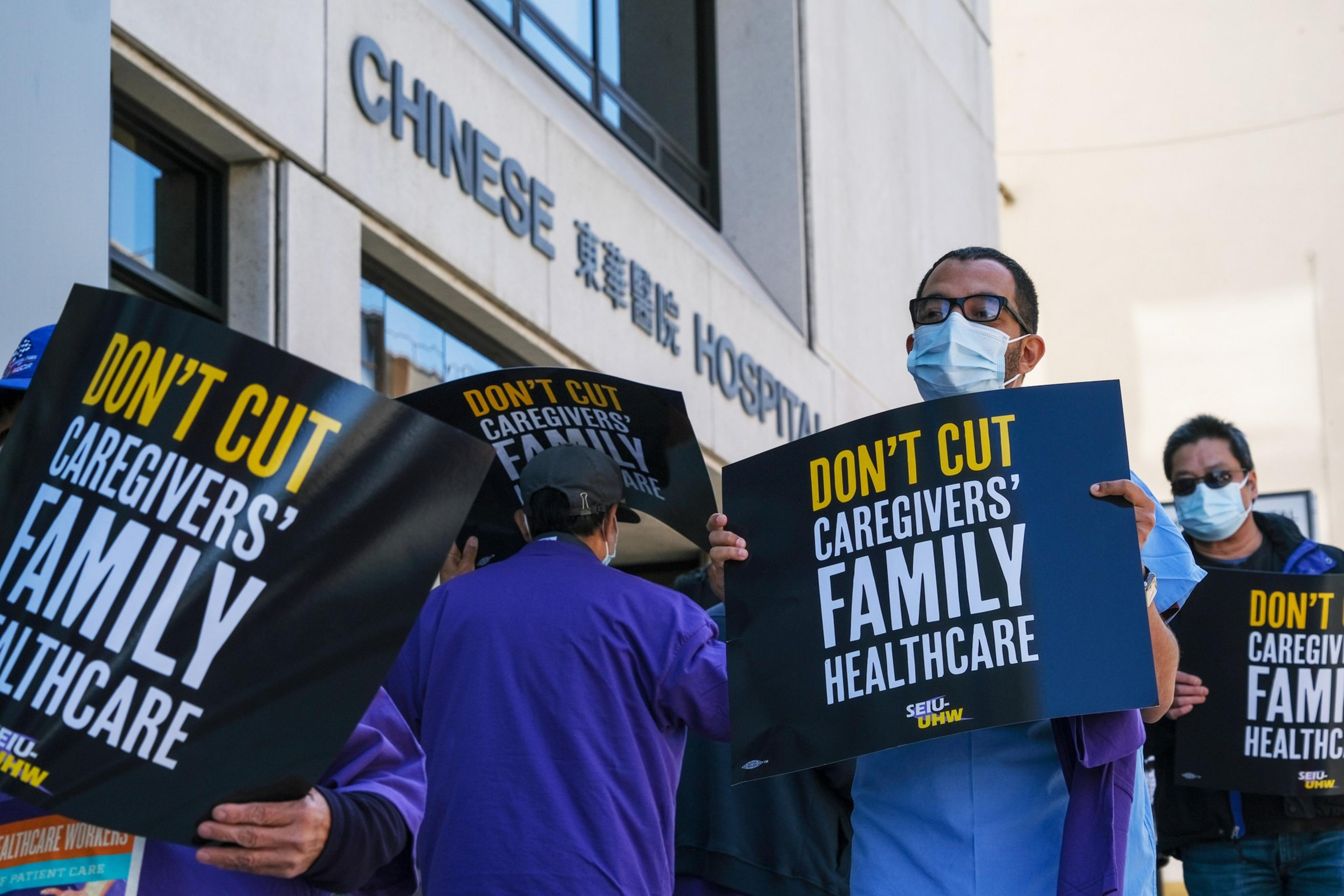People wearing masks hold signs reading "Don't cut caregivers' family healthcare" outside a building labeled Chinese Hospital.