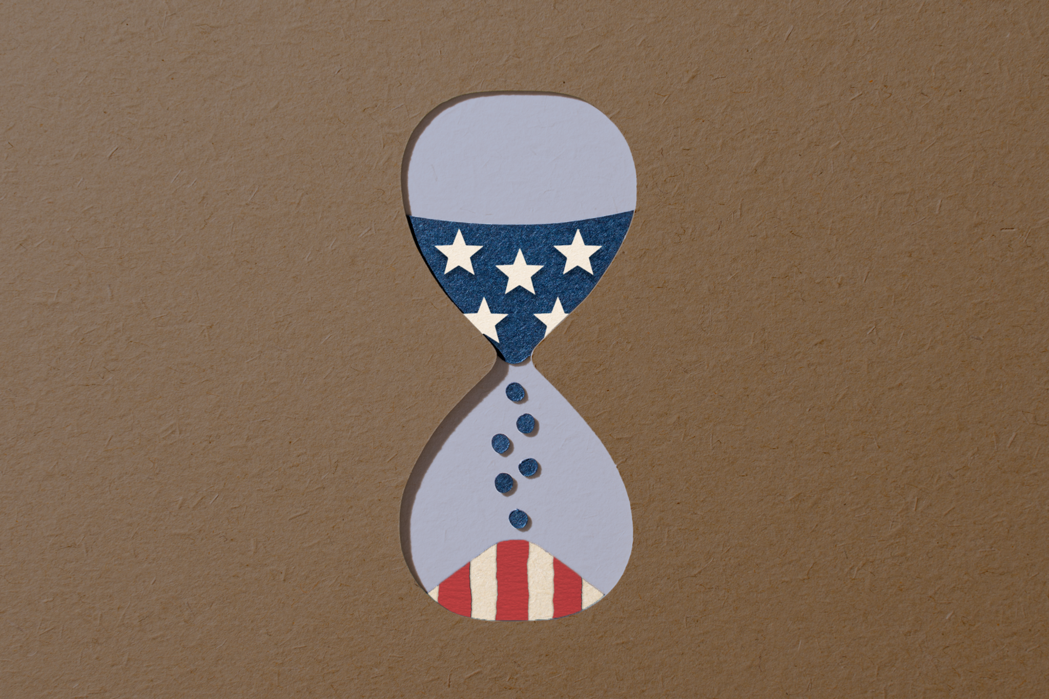 An hourglass with a U.S. flag pattern: stars on a blue background in the top half, and red and white stripes in the bottom half, set on a brown surface.