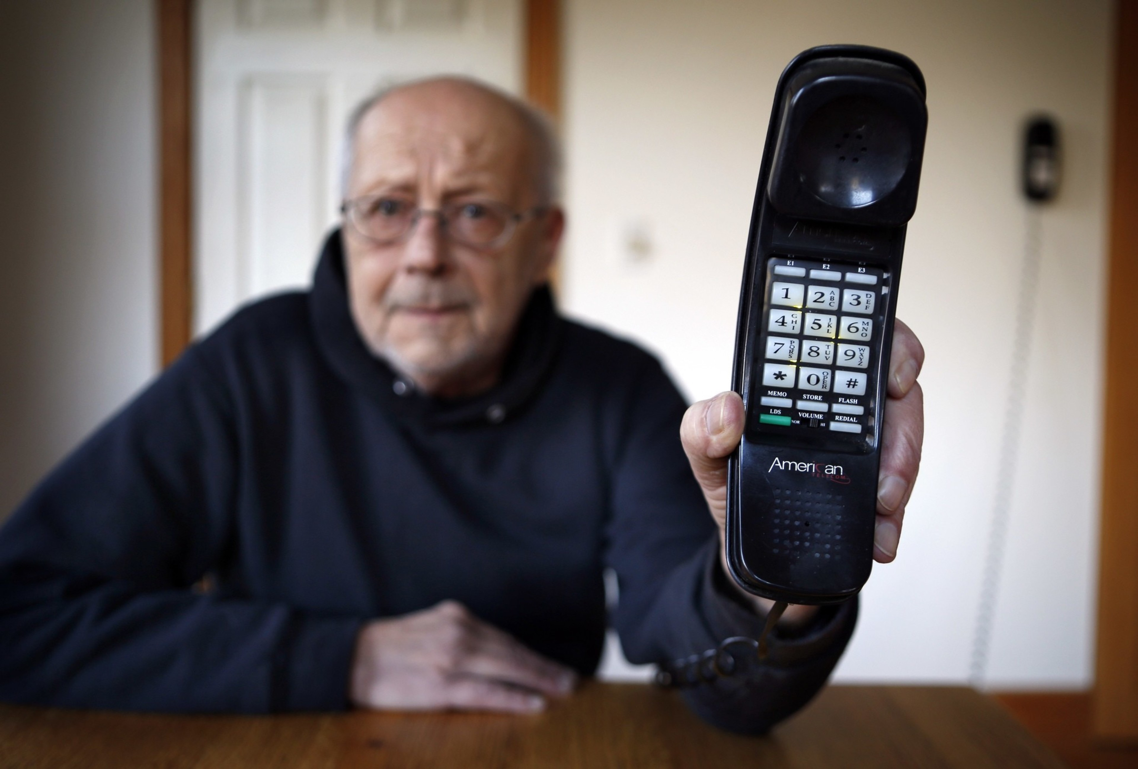 An elderly man holding out a large-buttoned landline phone, with a focused look.