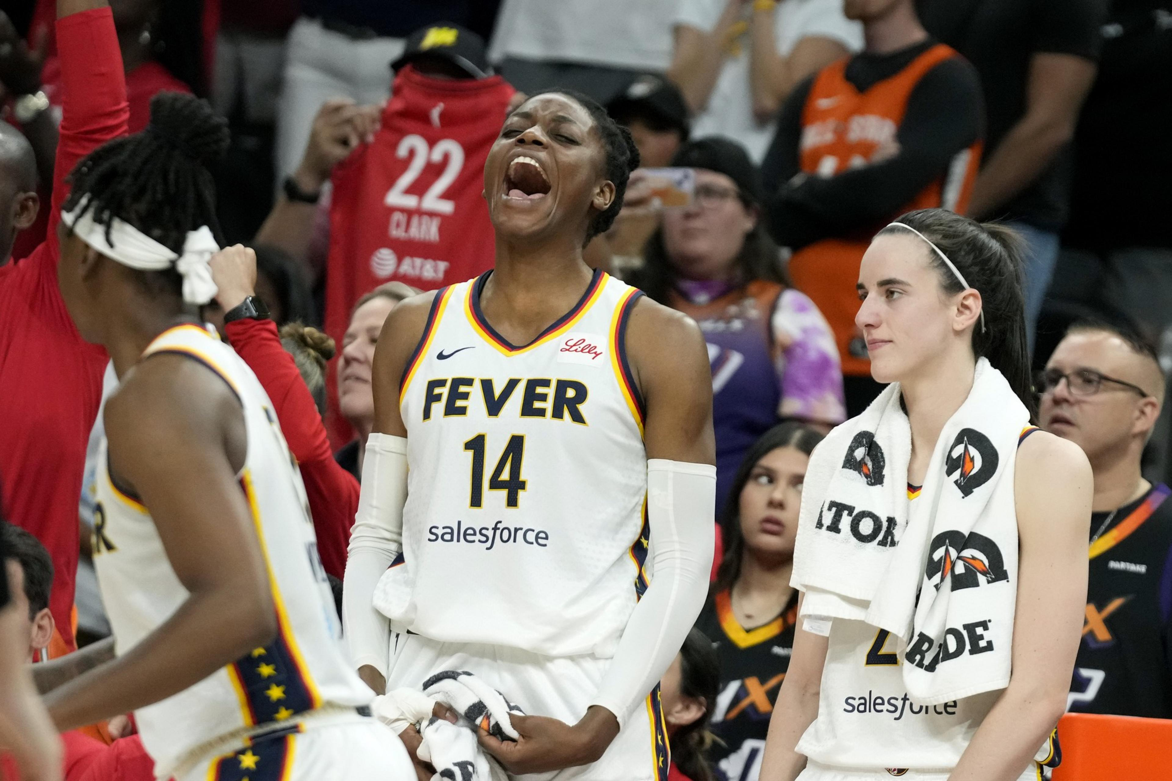 A basketball player wearing a &quot;Fever&quot; jersey shouts excitedly on the court, surrounded by teammates and fans in the background, creating an energetic atmosphere.