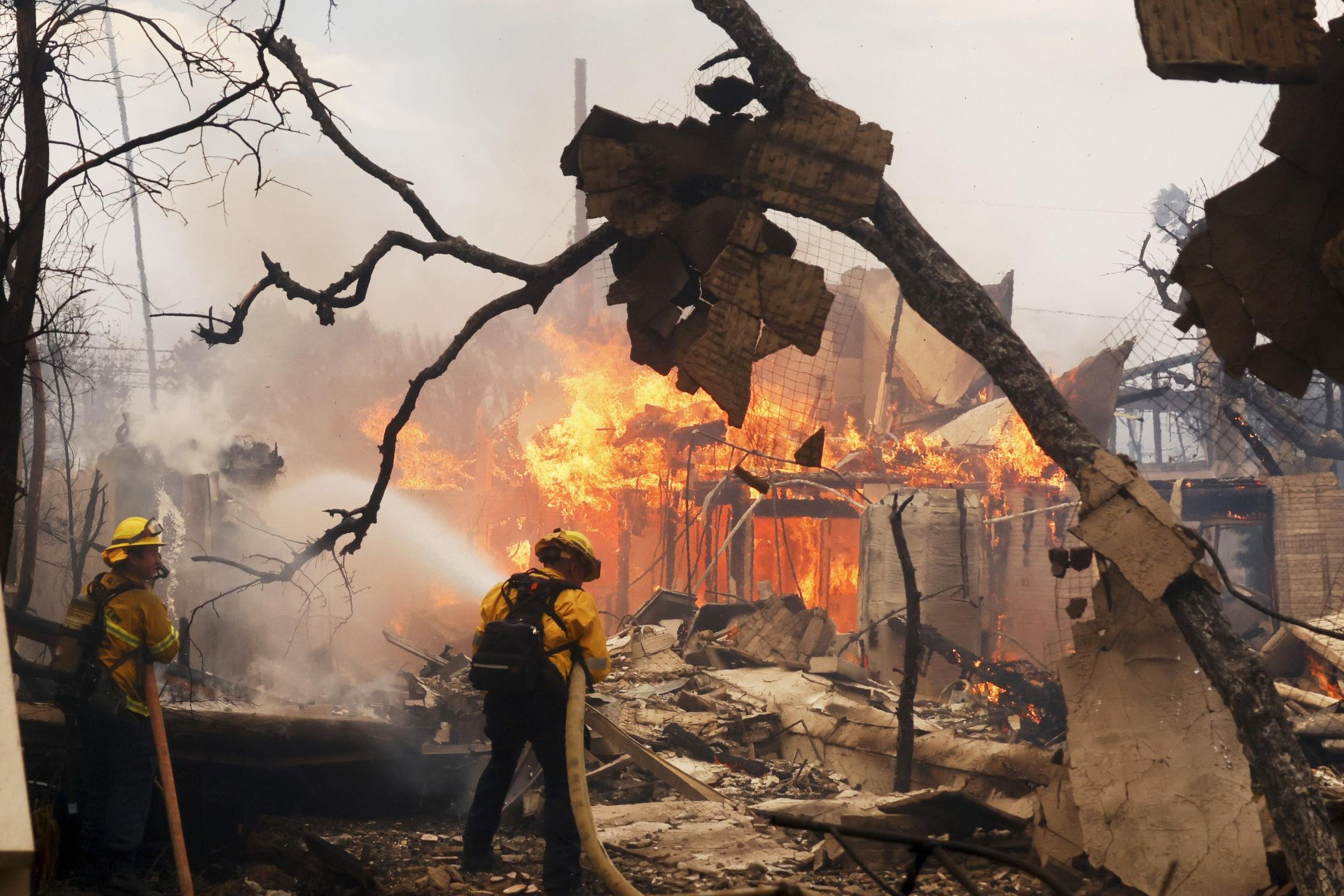 Firefighters in yellow gear are battling a large blaze engulfing a building, surrounded by debris and charred trees, using hoses to spray water.
