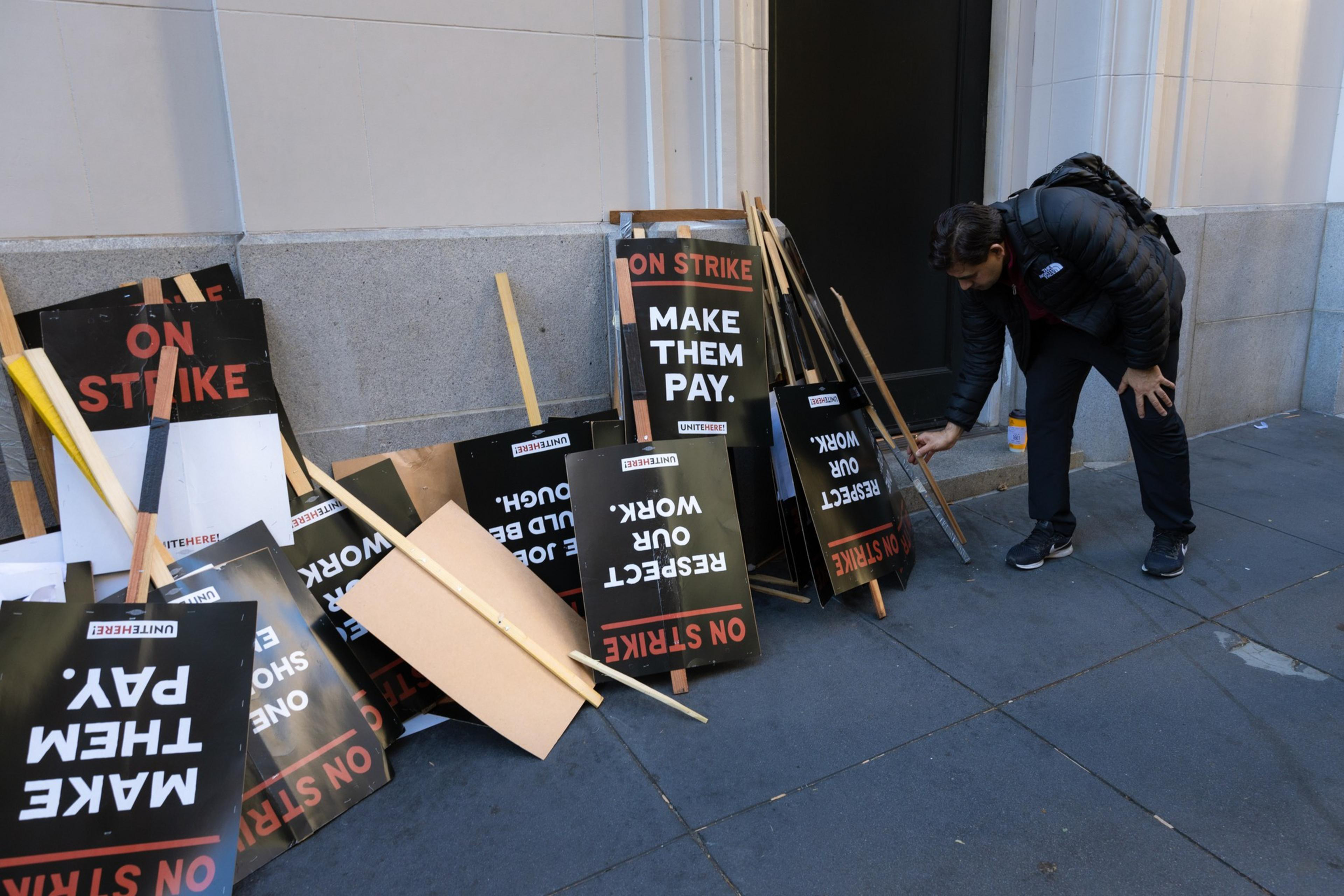 A man with a backpack picks up a protest sign near a pile of other signs leaning against a building. The signs read, &quot;ON STRIKE,&quot; &quot;MAKE THEM PAY,&quot; and &quot;RESPECT OUR WORK.&quot;