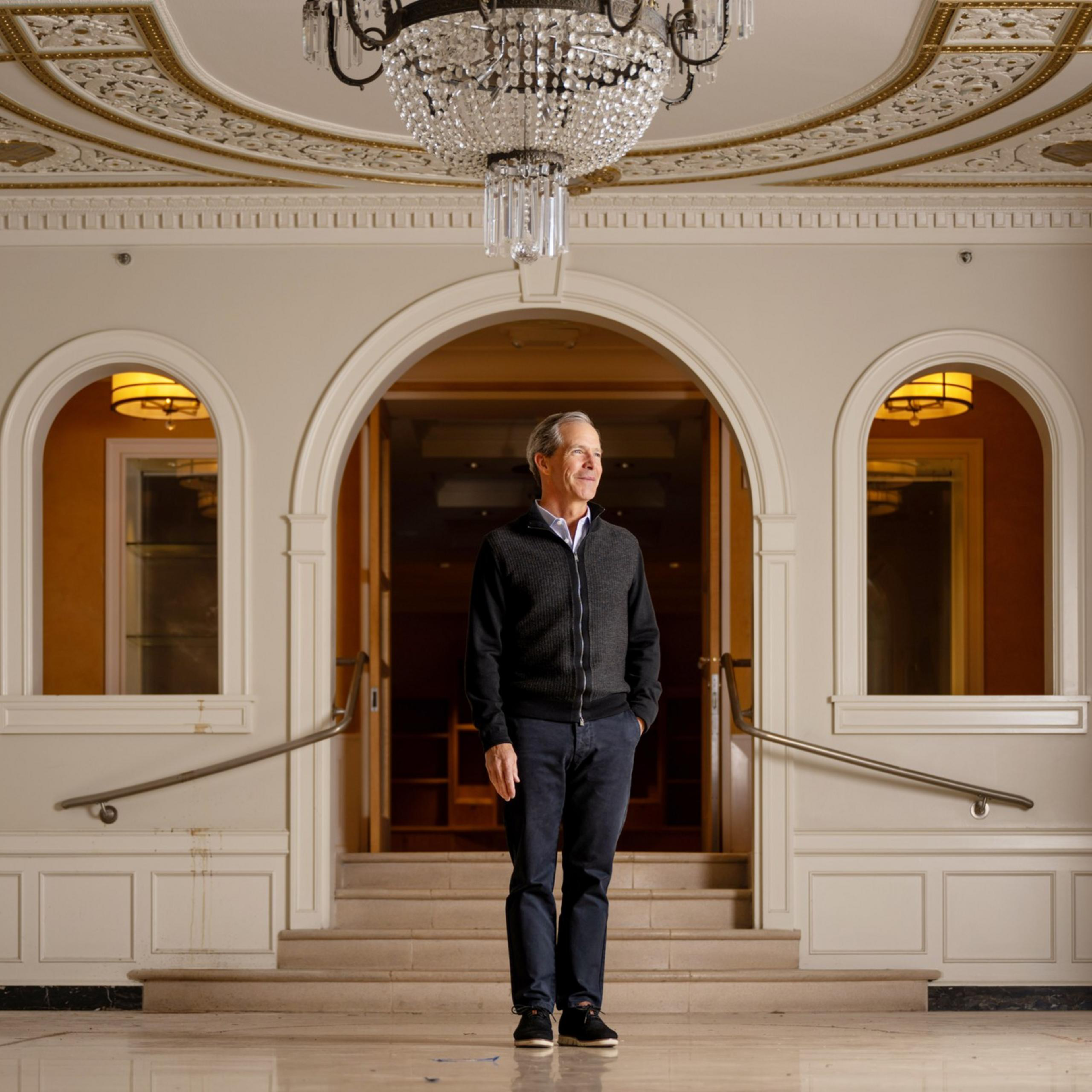 A person in a dark sweater stands in a brightly lit, ornate room with white walls, arched doorways, a chandelier, and decorative ceiling details.