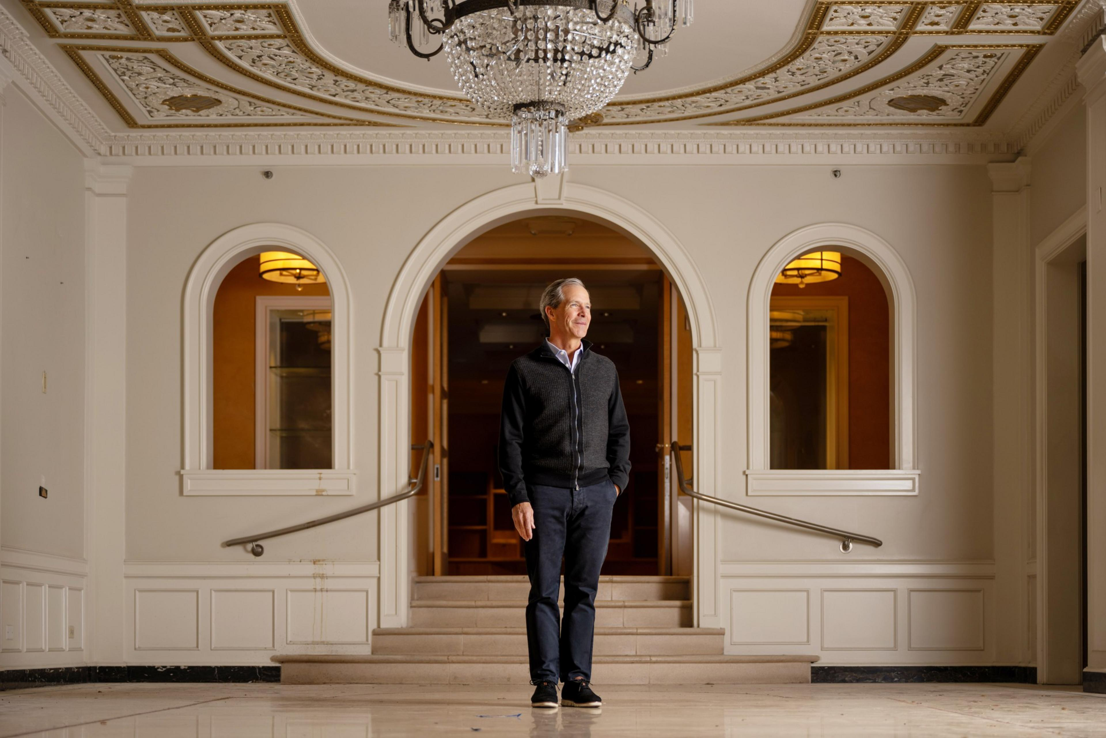A person in a dark sweater stands in a brightly lit, ornate room with white walls, arched doorways, a chandelier, and decorative ceiling details.