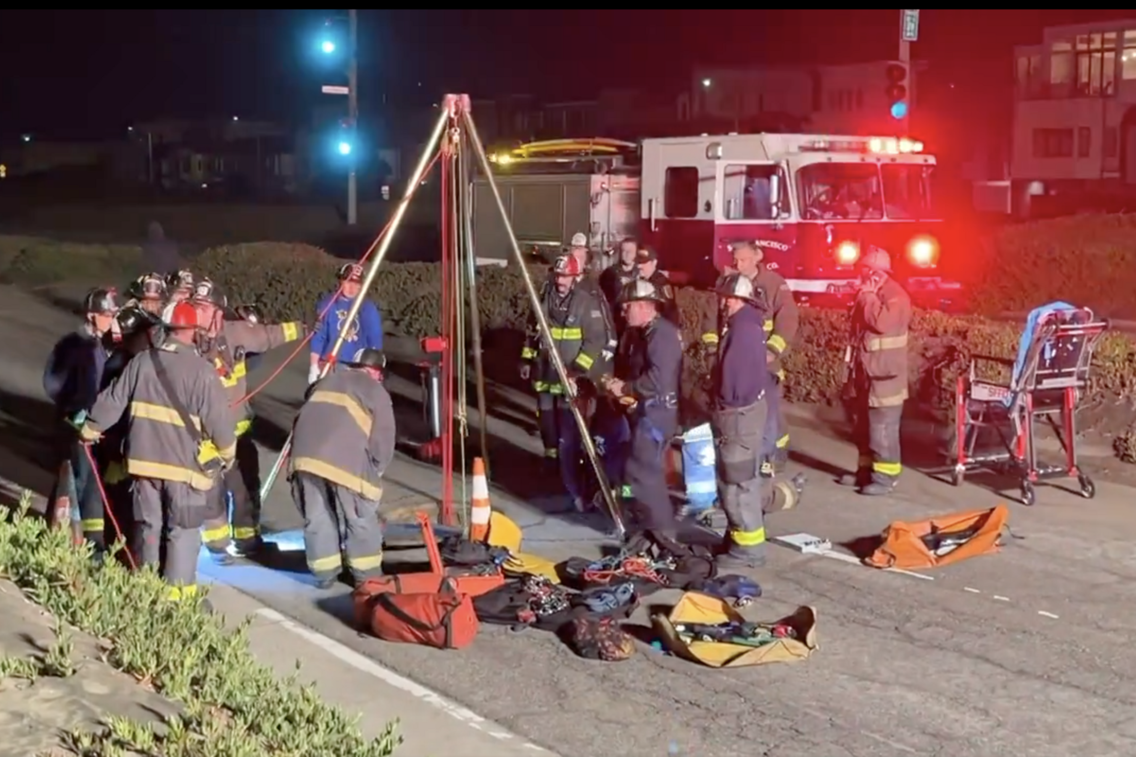 Firefighters are gathered around a tripod setup with ropes and equipment on the roadside at night, illuminated by emergency vehicle lights in the background.