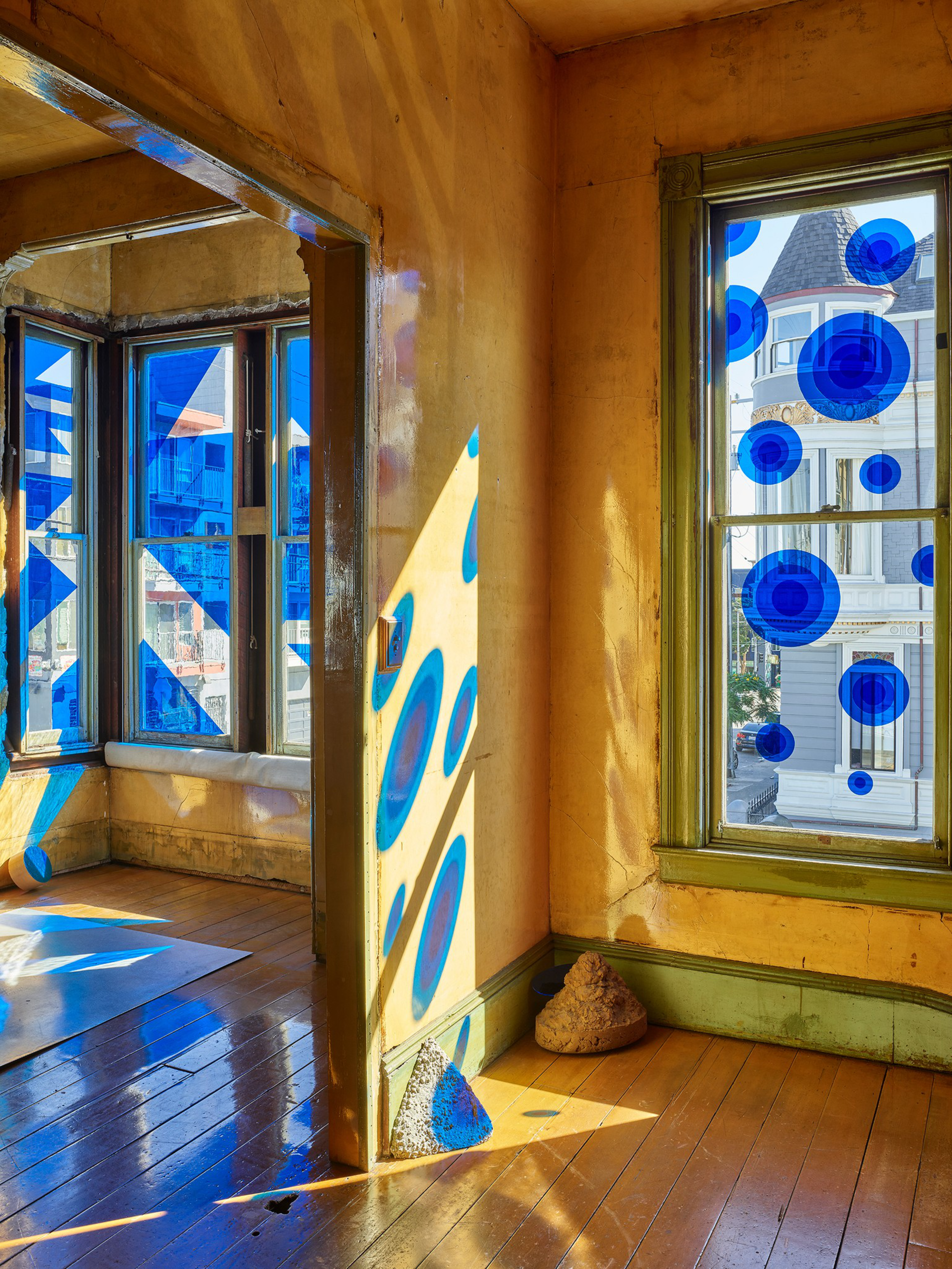 Sunlight streams through windows decorated with blue circular and geometric patterns, casting vibrant blue shapes on the warm, aged walls and wooden floor.