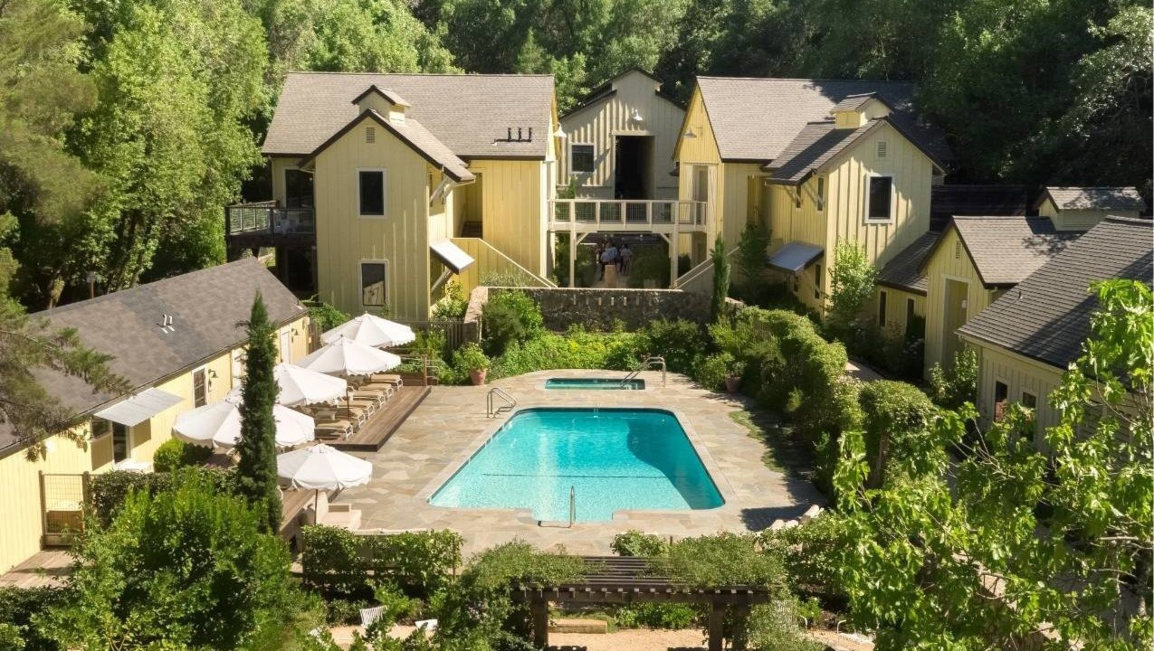 The image shows a charming estate with several yellow buildings surrounding a central swimming pool, nestled in lush greenery.