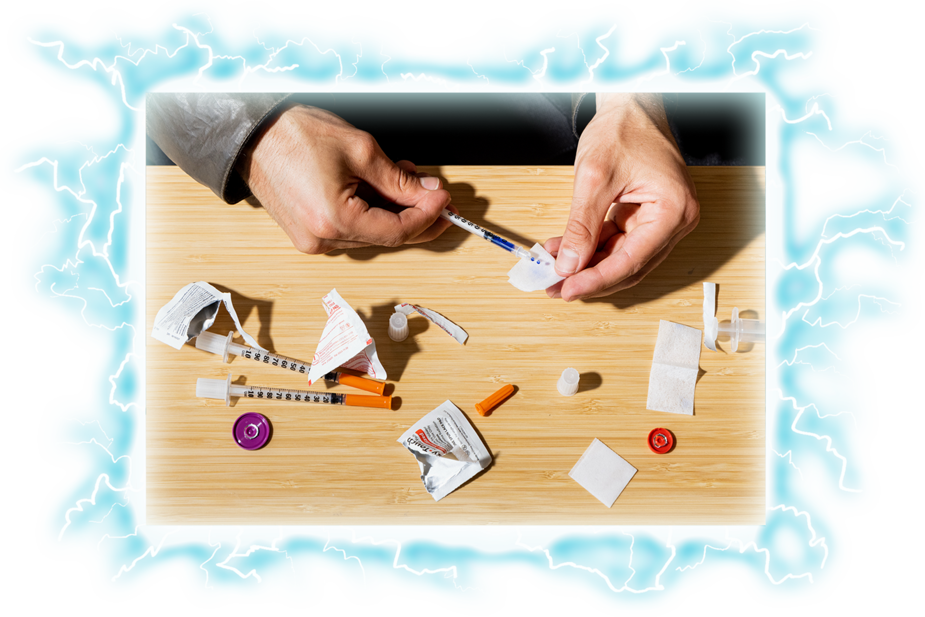 Two hands prepare a syringe on a wooden surface scattered with syringes, caps, alcohol wipes, and torn packaging.