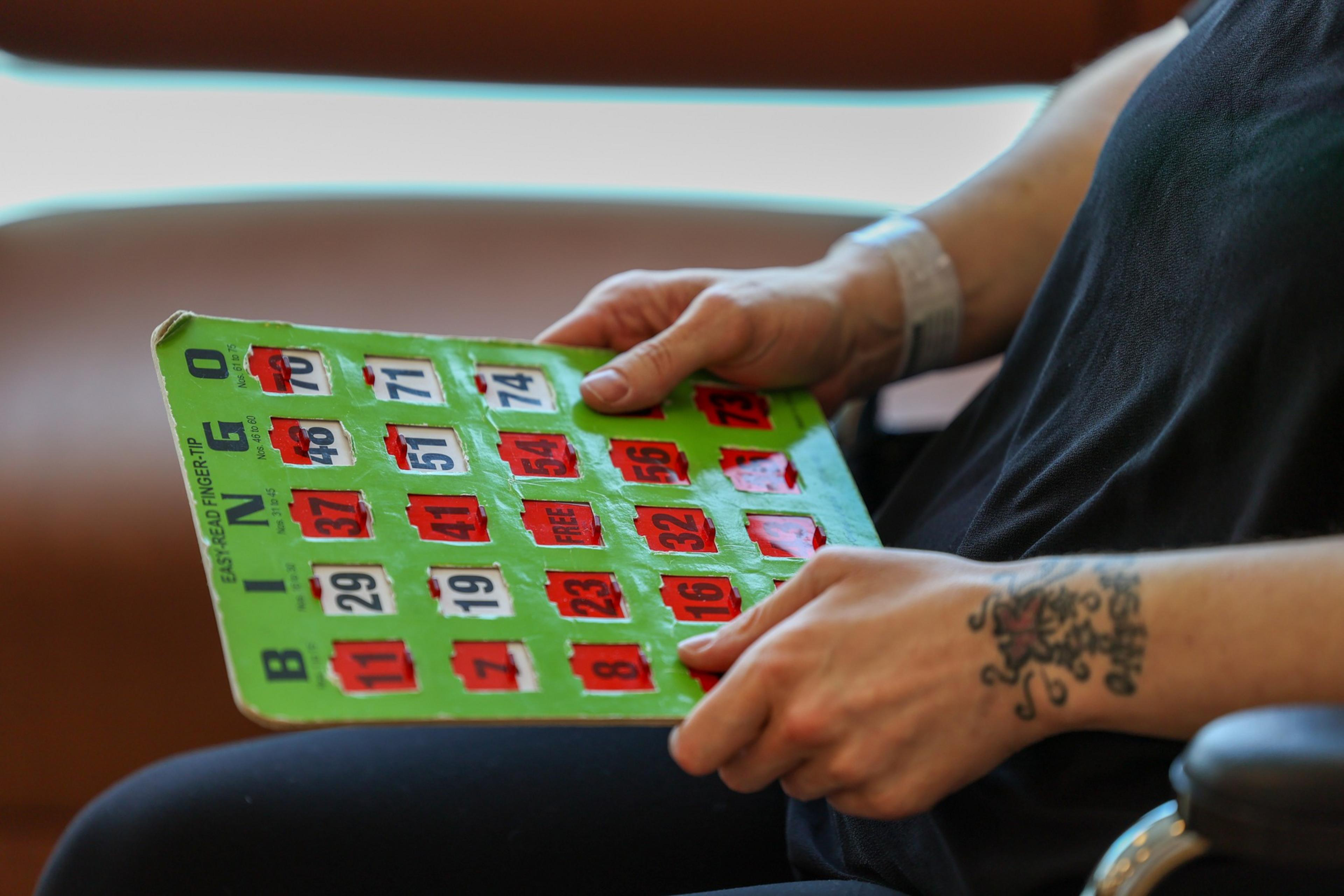 A person with a tattoo on their right wrist holds a green bingo card with many numbers marked off in red.