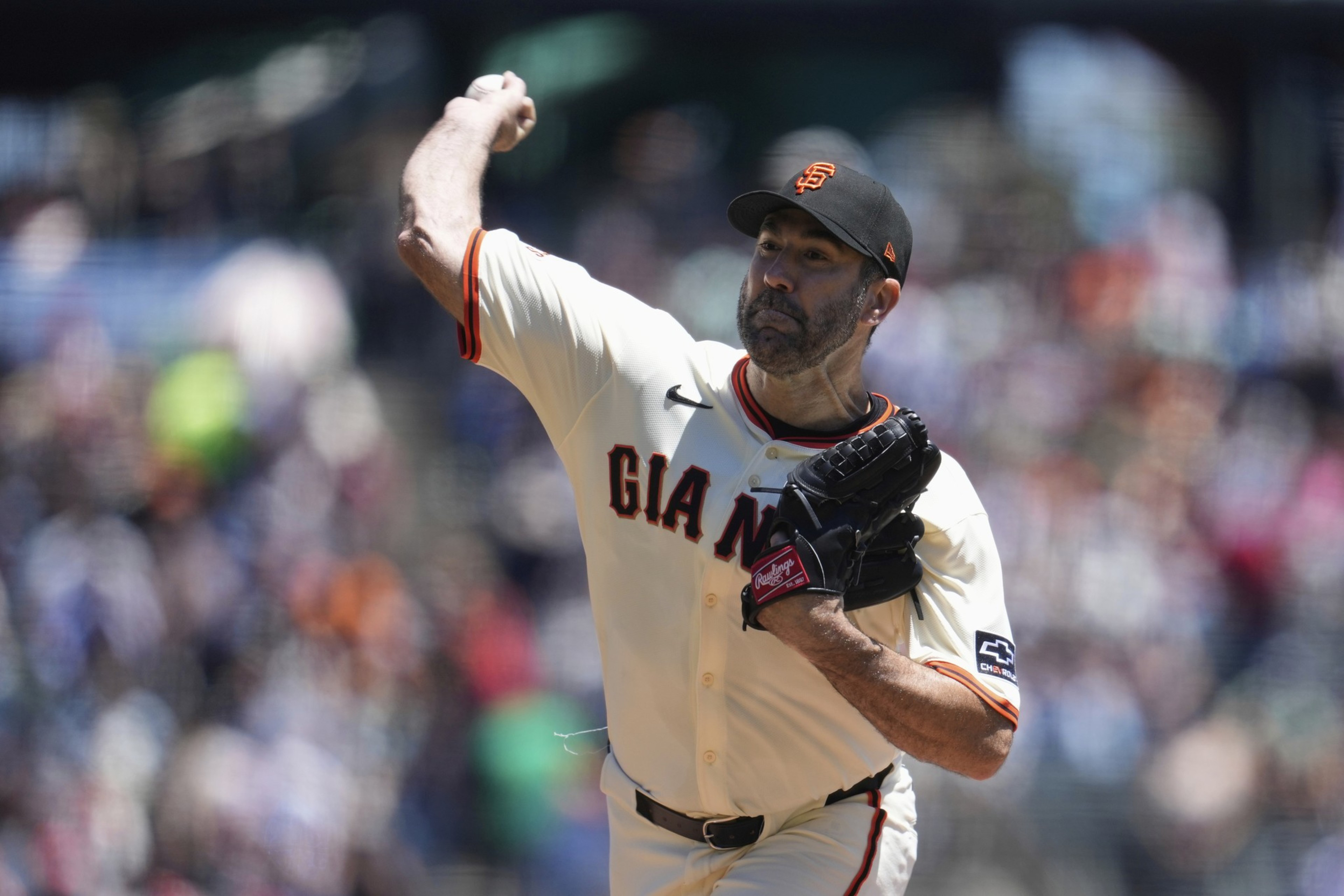 A baseball player in a white jersey with "GIANTS" is pitching. He wears a black cap with an orange logo, and a black glove, against a blurred stadium backdrop.