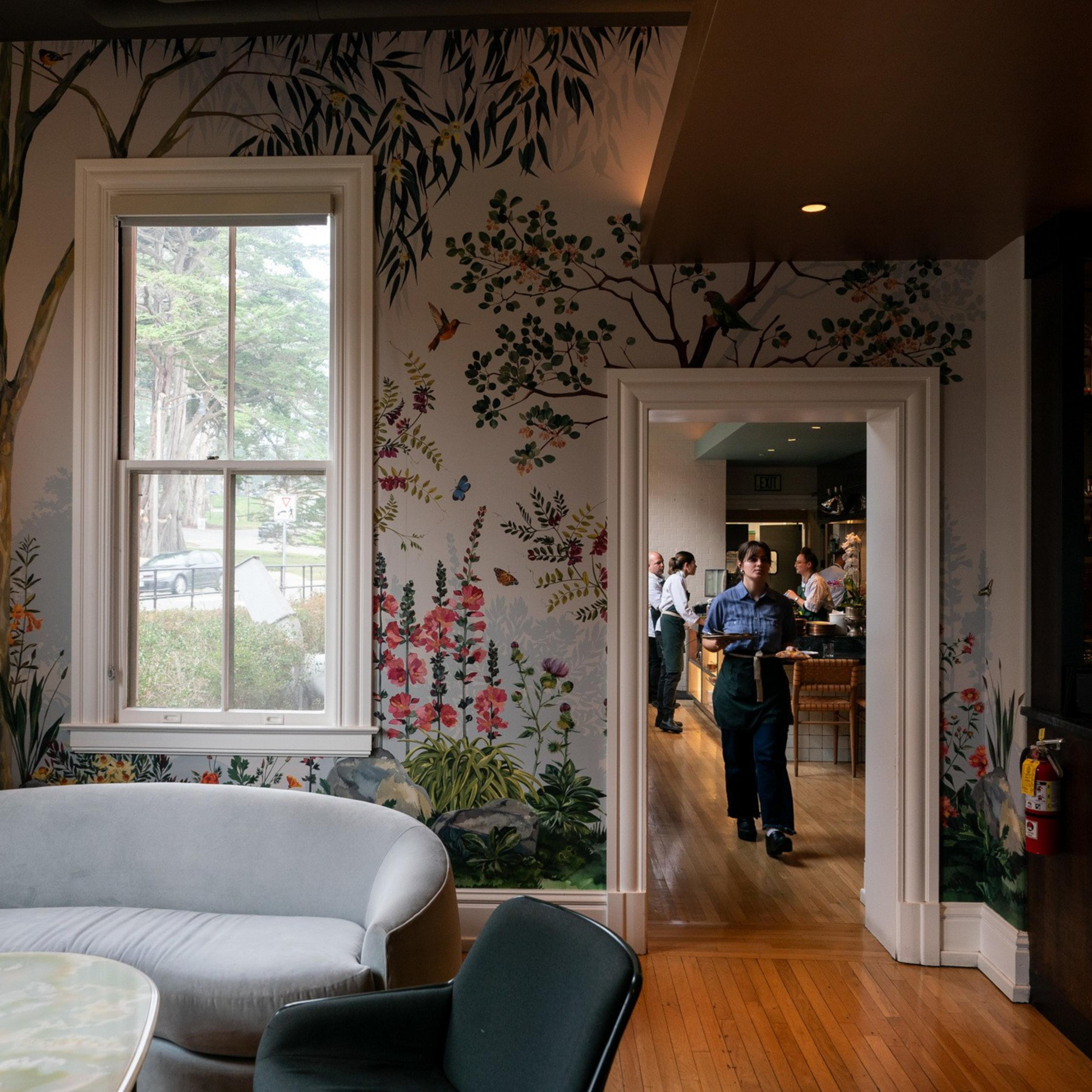 A cozy room with floral and bird murals, featuring a gray sofa and a person carrying drinks from an adjacent room. A bar with bottles is to the right.