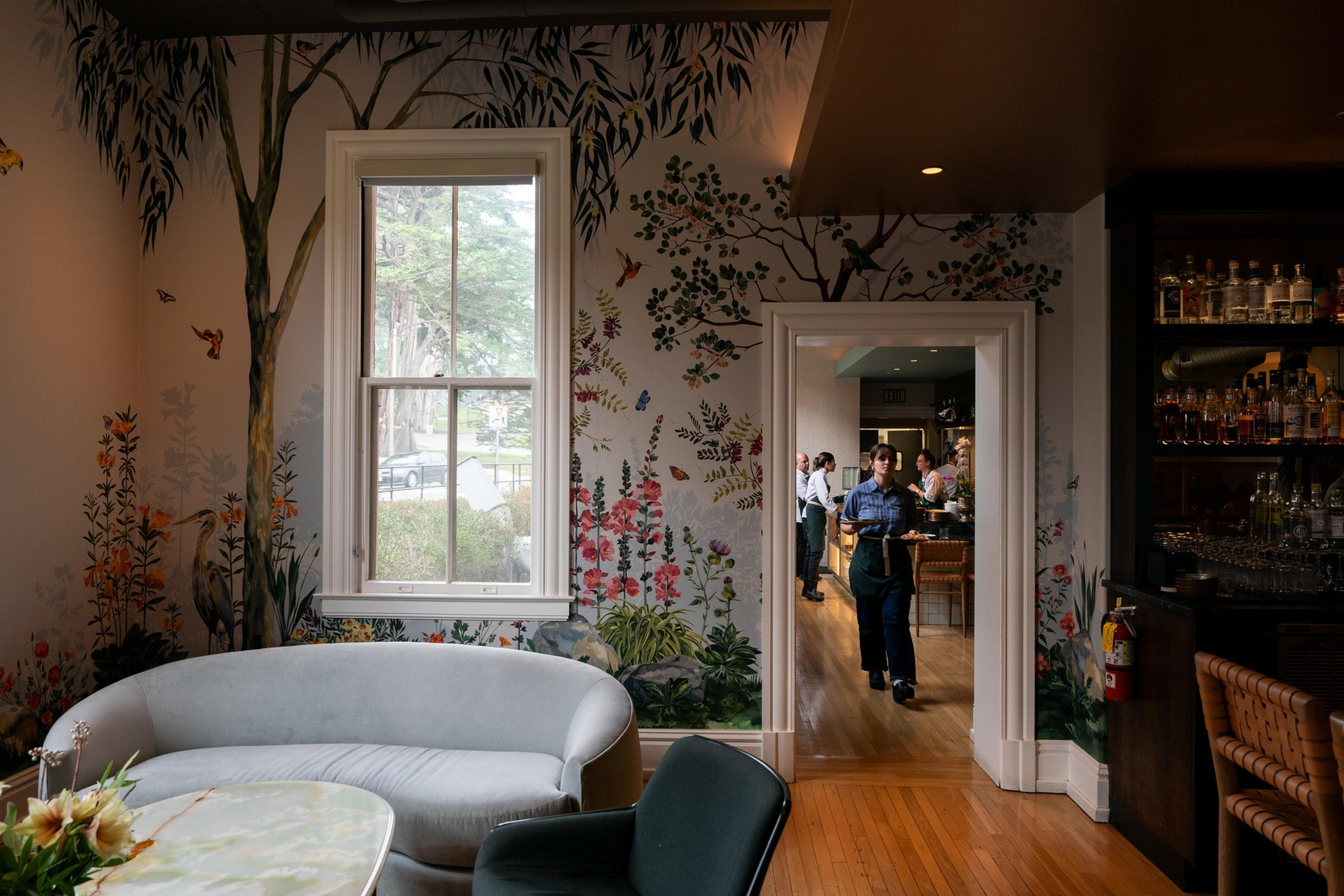 A cozy room with floral and bird murals, featuring a gray sofa and a person carrying drinks from an adjacent room. A bar with bottles is to the right.