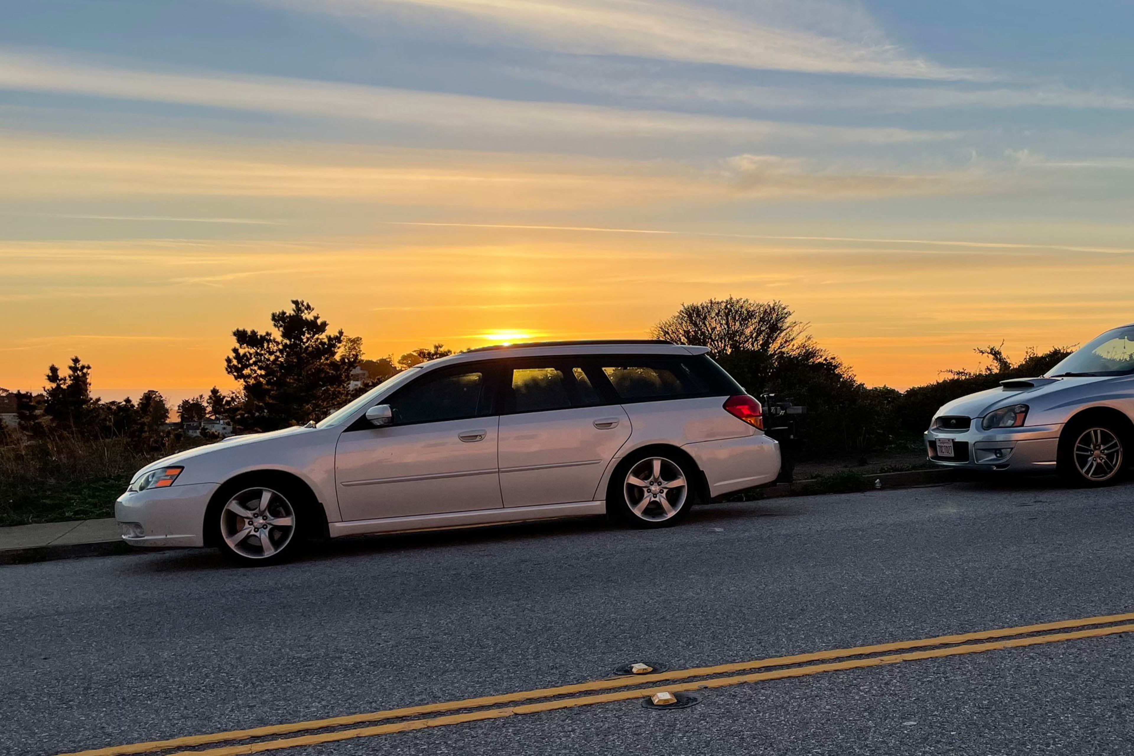 A silver station wagon and a silver sedan are parked along a quiet road with a sunset and trees in the background.