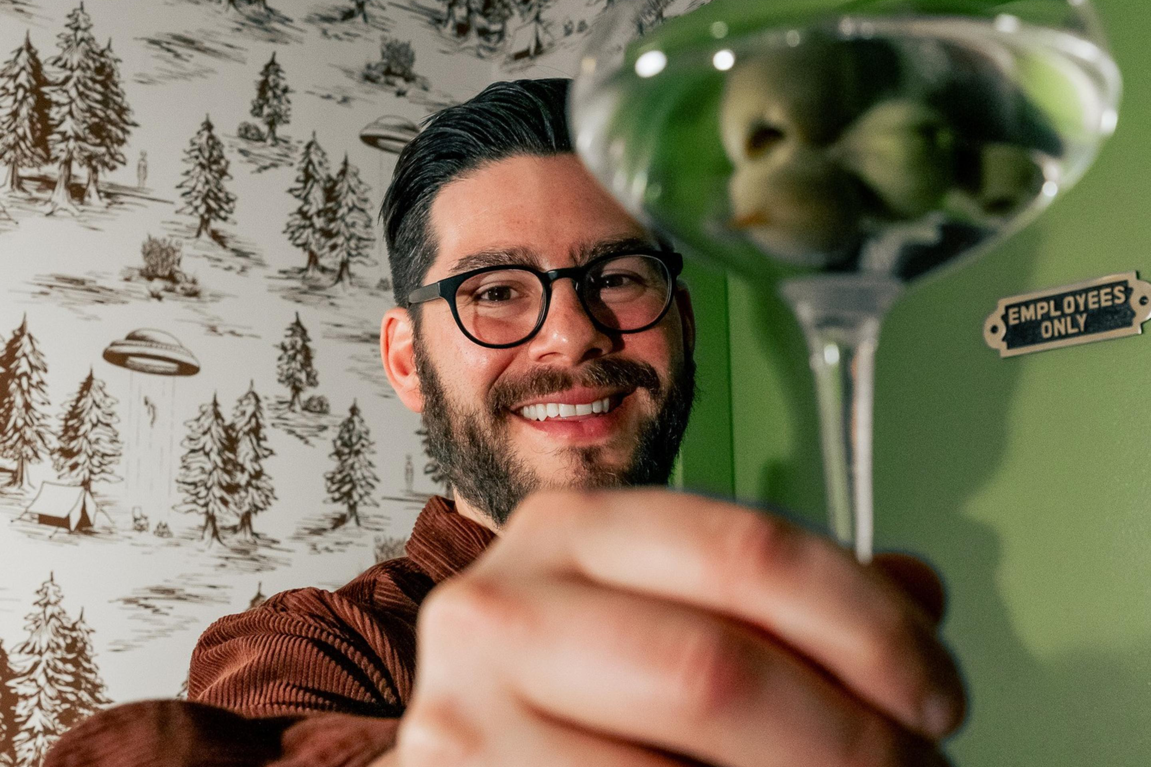 A person wearing glasses and a brown shirt smiles at the camera, holding up a martini glass with olives. The background features a forest-themed wallpaper.