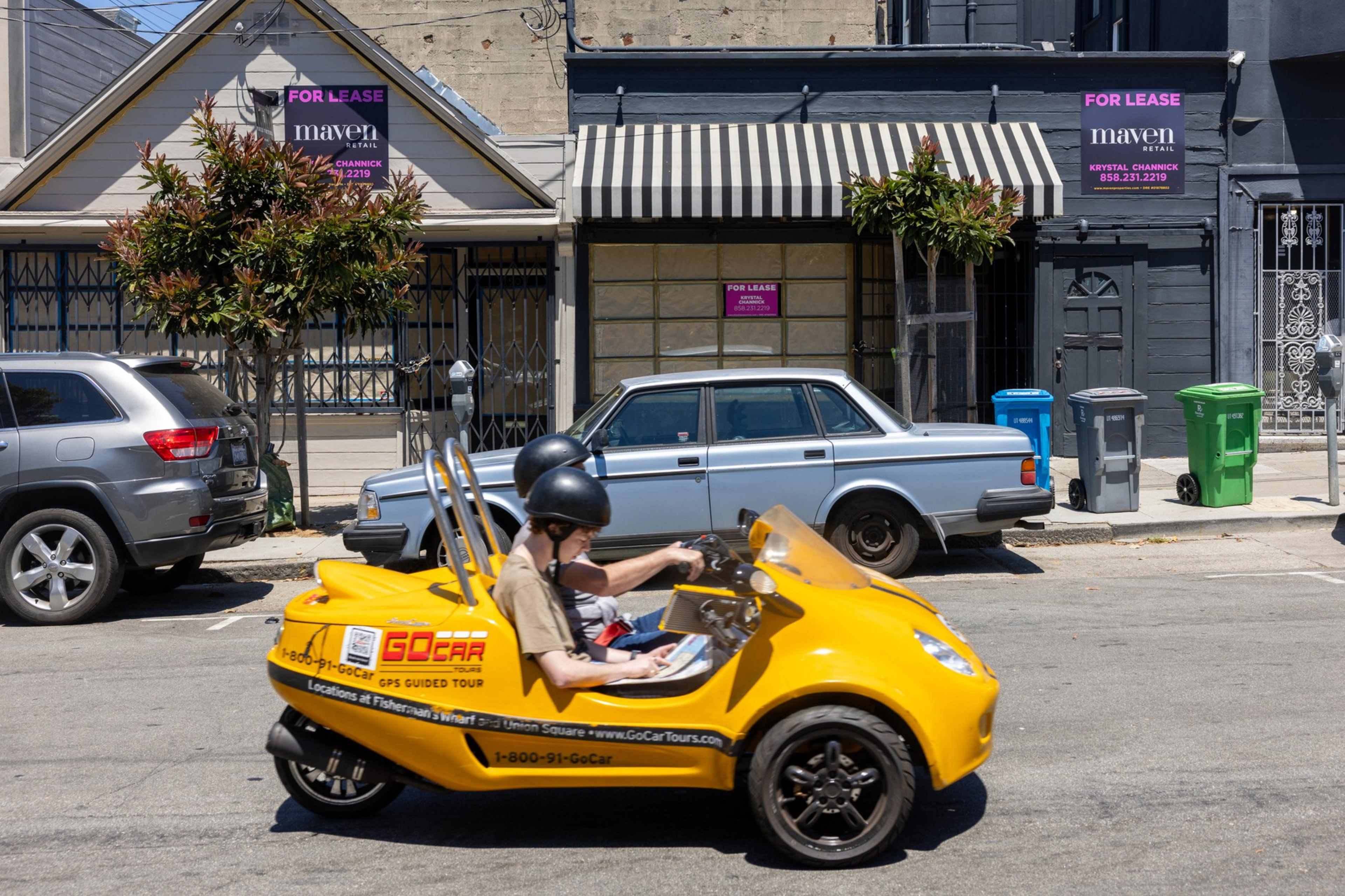 A yellow GoCar with two helmeted passengers drives past parked cars and two "For Lease" signs on a street with gray buildings and a striped awning.