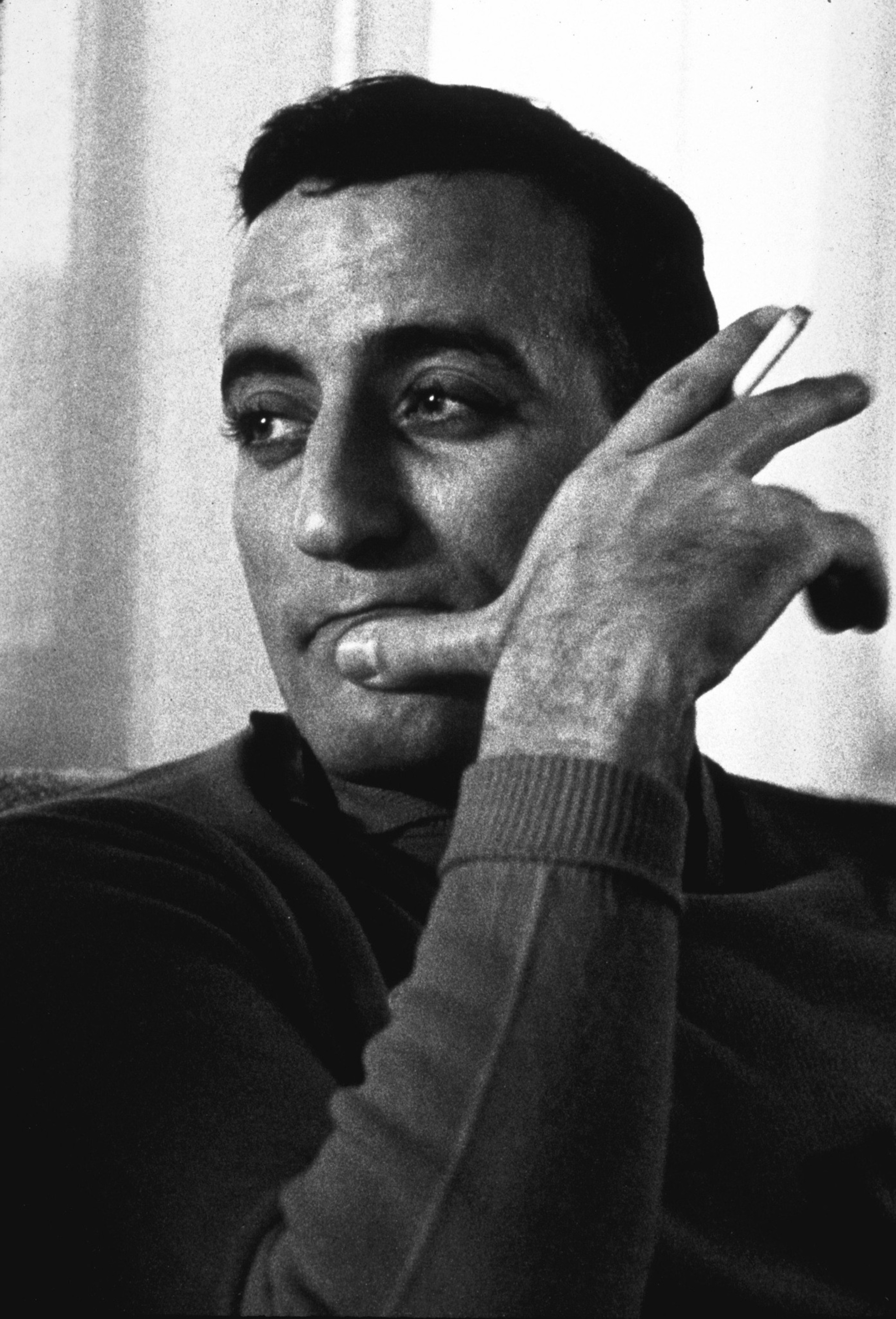 A man with dark hair and a contemplative expression holds a cigarette near his face, looking off to the side in a black-and-white photo.