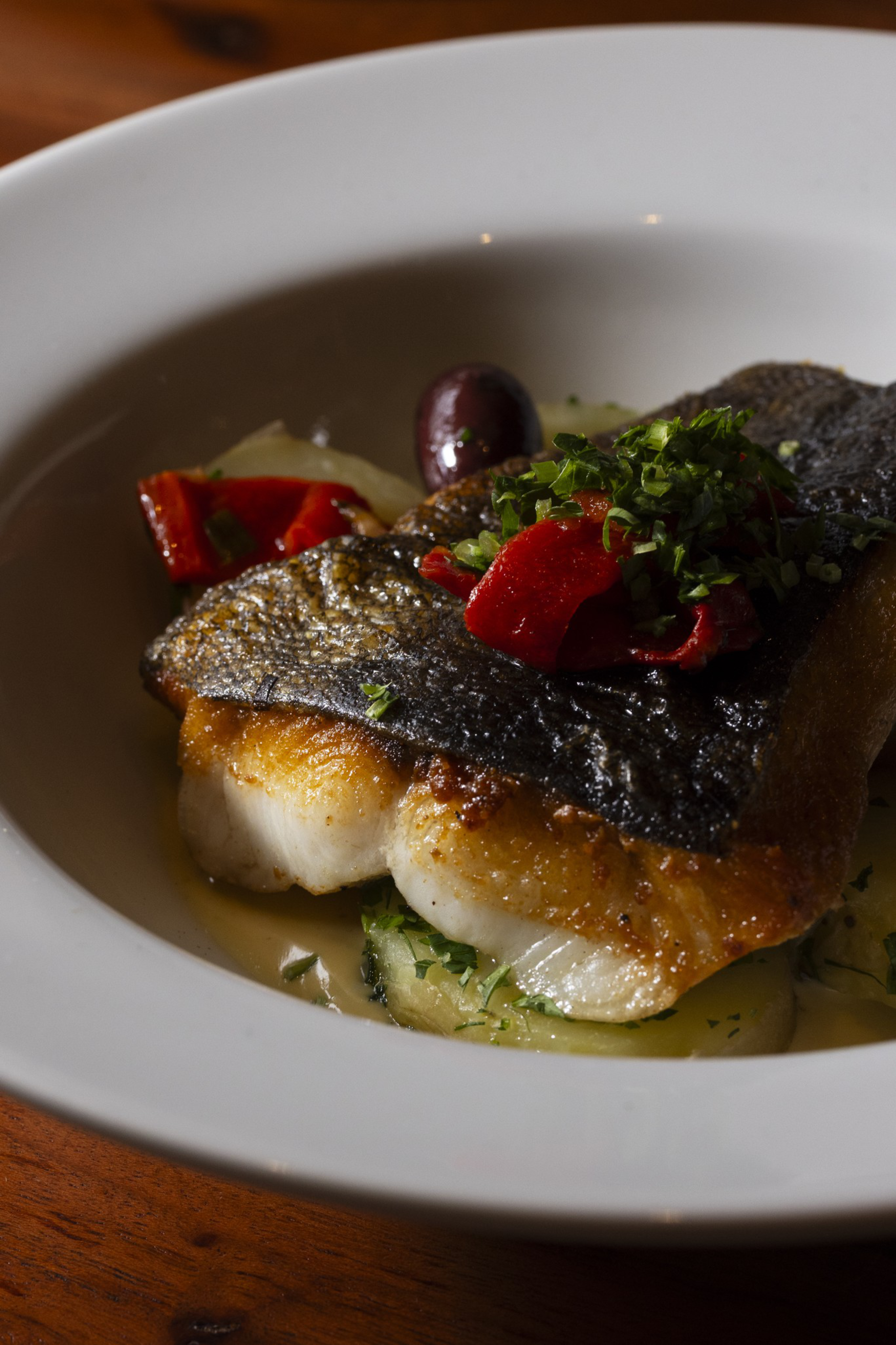 A seared fish fillet with crispy skin sits on a bed of sliced potatoes, garnished with chopped herbs, red peppers, and a black olive.