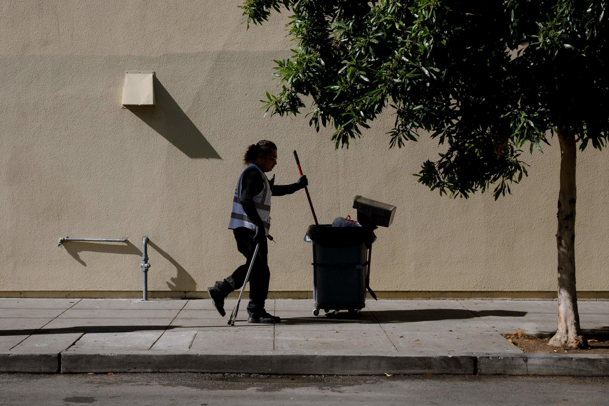 A person wearing a reflective vest pushes a trash bin and holds a broom while walking on a sidewalk next to a beige wall and a tree.