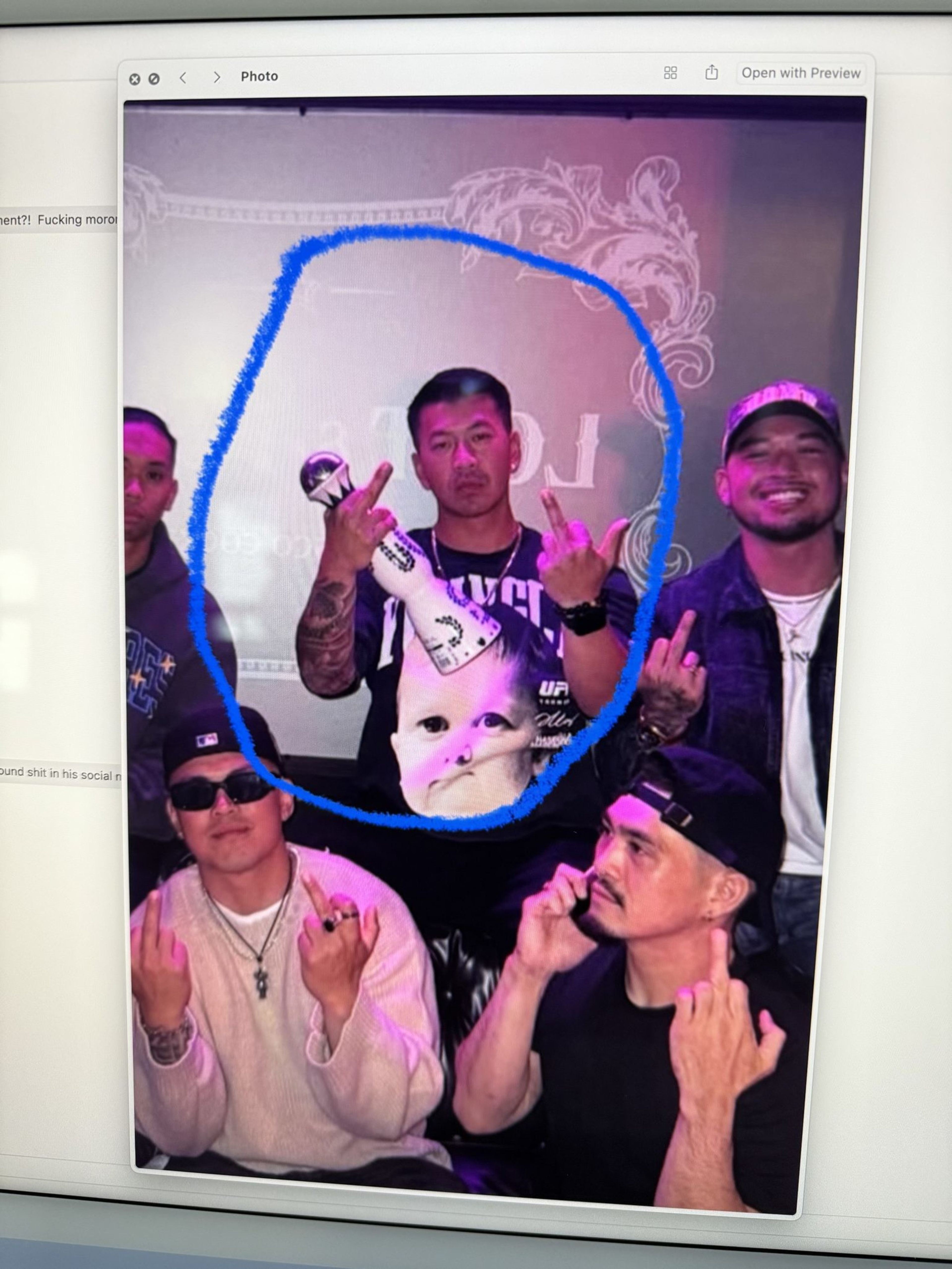 Five men are posing, making rude gestures. One in the center holds a statue, wearing a shirt with a baby face. The atmosphere is casual with bright lighting.
