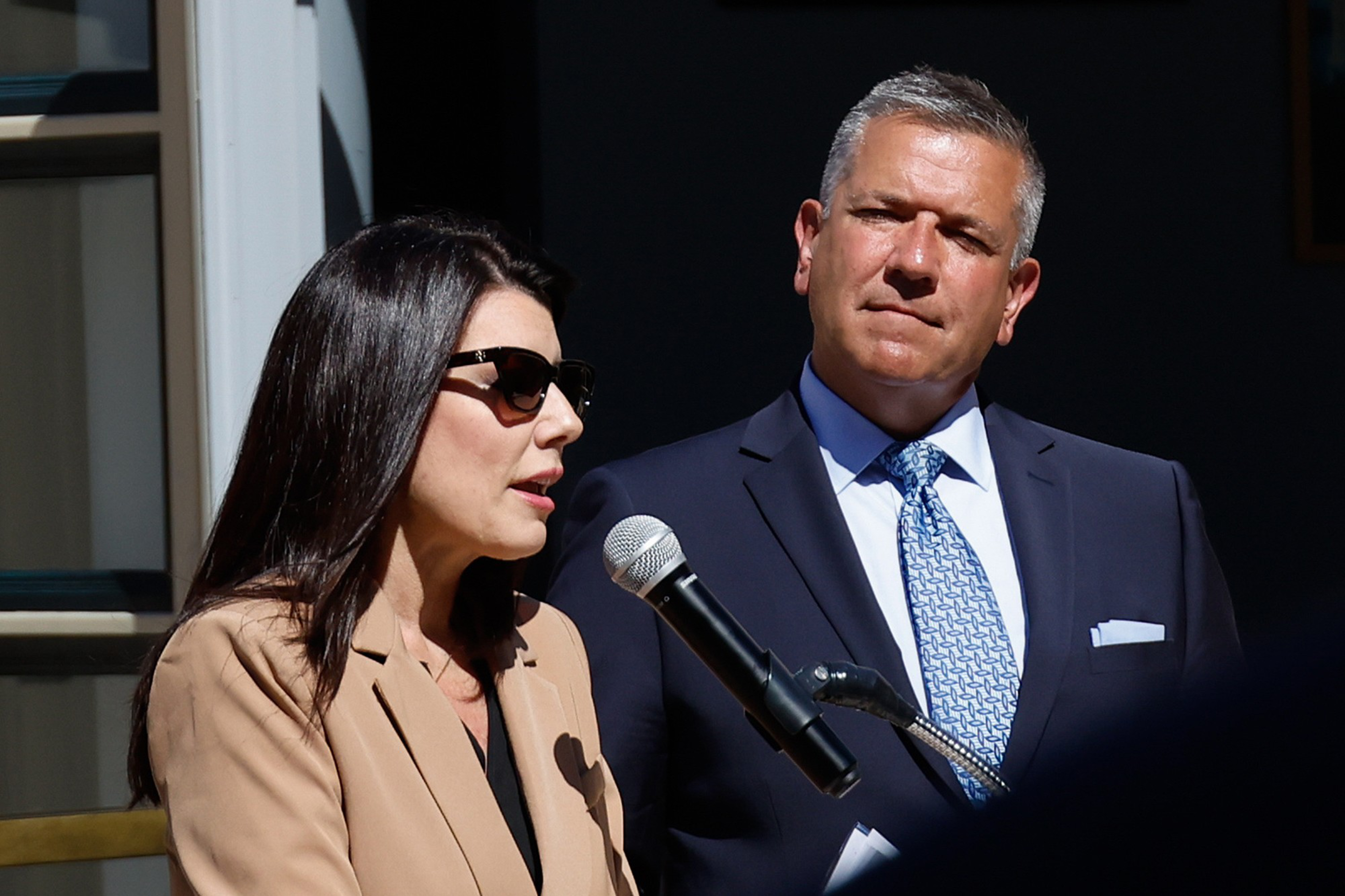 A woman wearing sunglasses and a tan blazer speaks into a microphone while a man in a blue suit and patterned tie listens attentively.