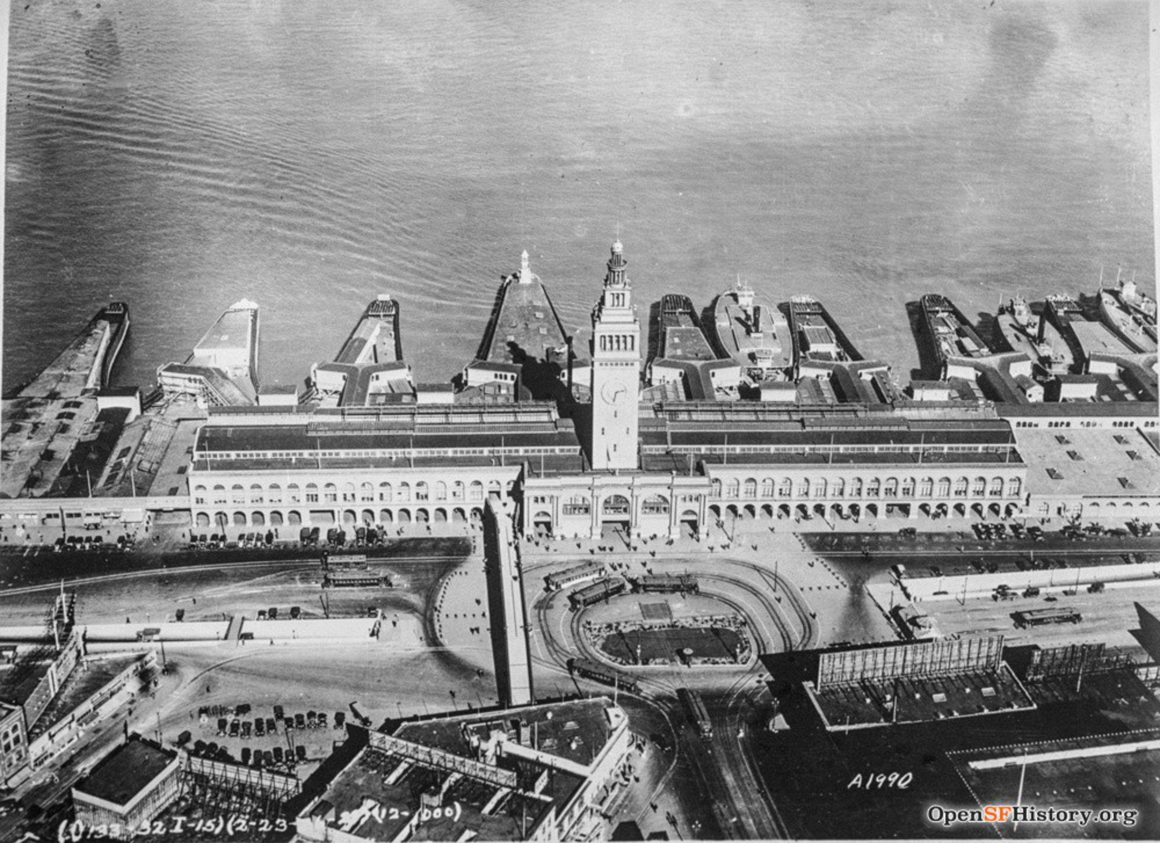 Aerial view of a large waterfront building with a tall clock tower, piers extending into the water, cars, and people around the plaza.