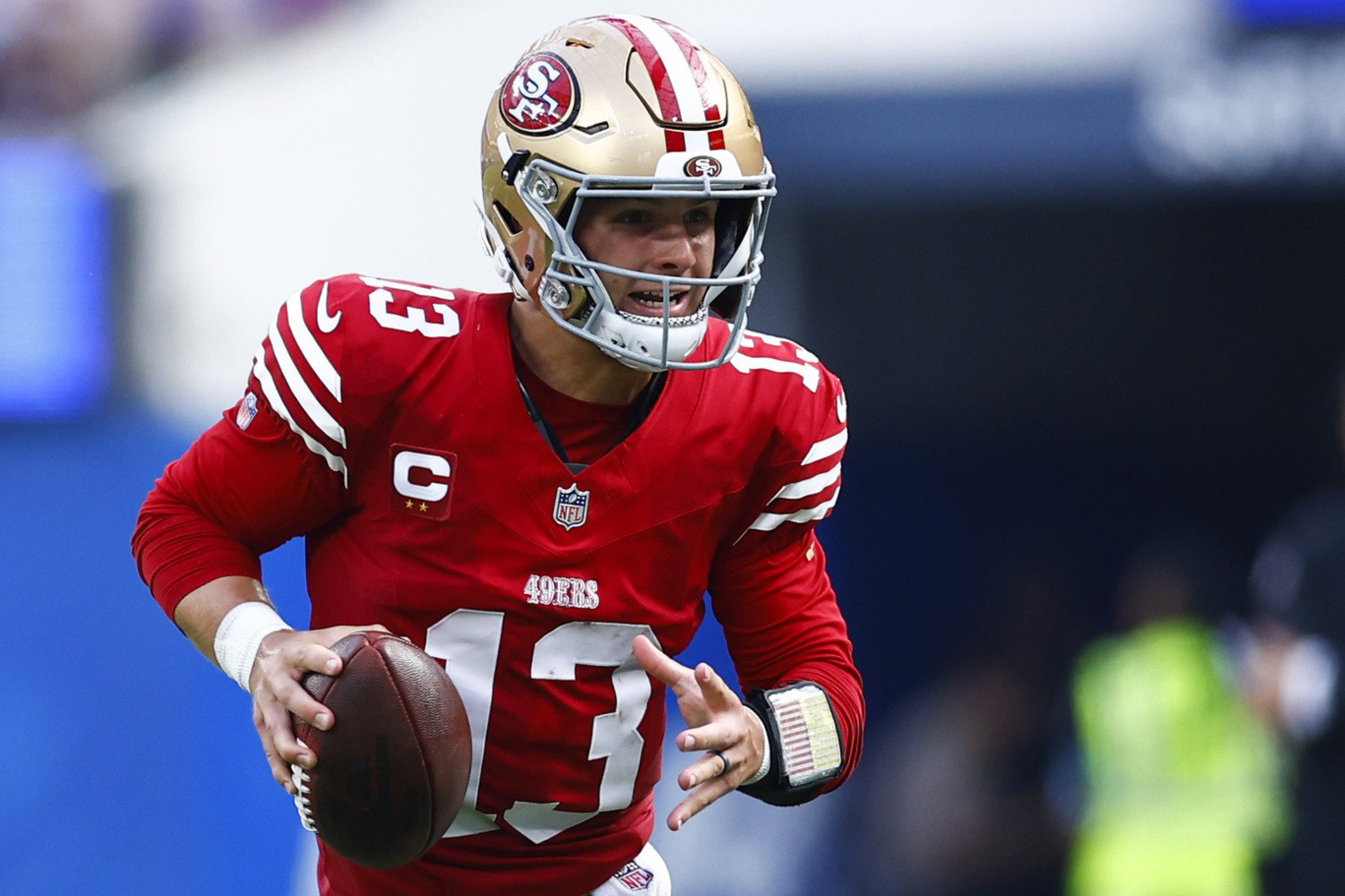 A football player in a red and gold uniform is running with the ball, looking ahead, likely to pass. The field is green turf, and the background is blurred.