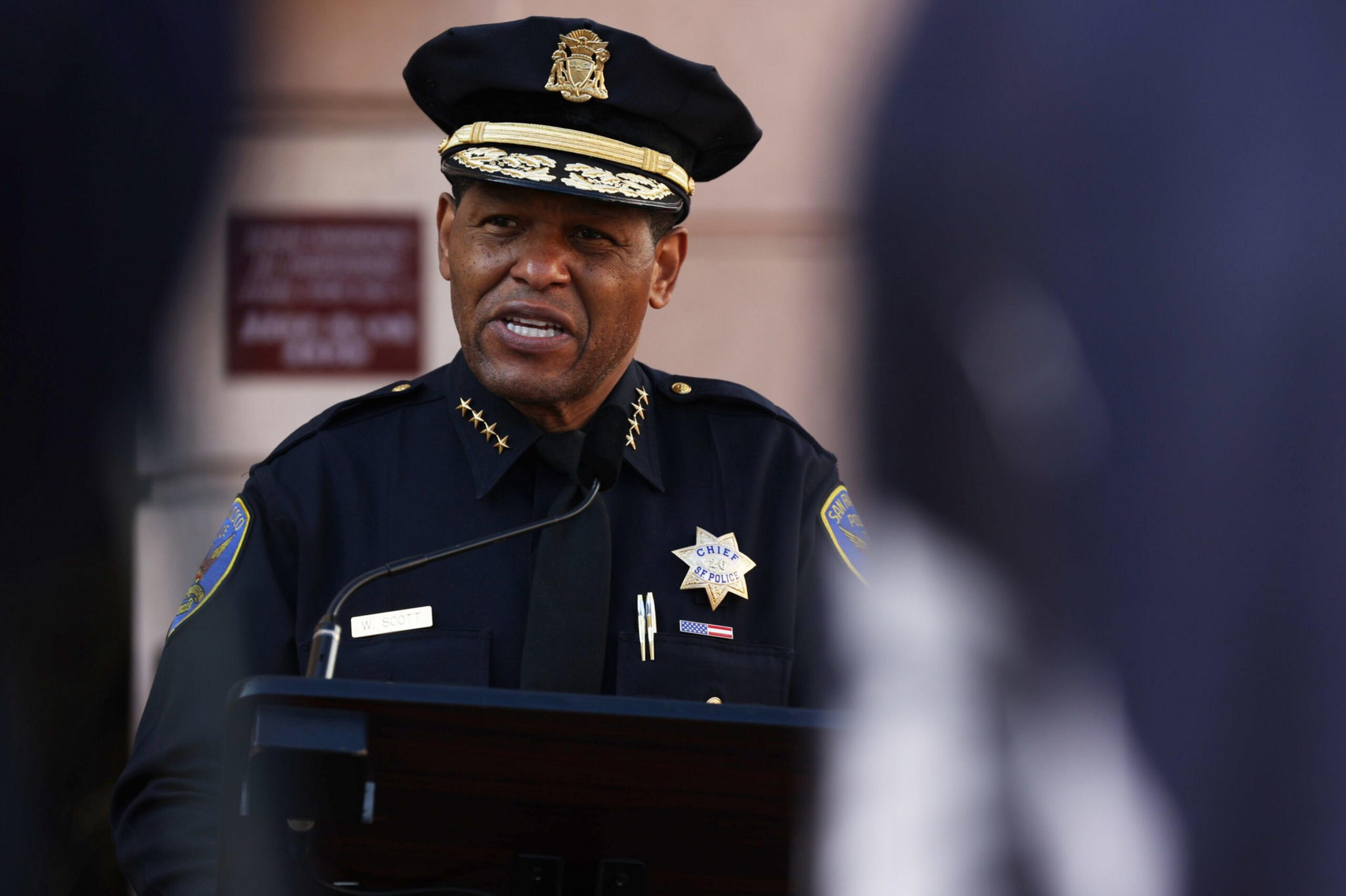 A police chief in uniform speaks at a podium with a microphone, framed by blurred figures in the foreground.
