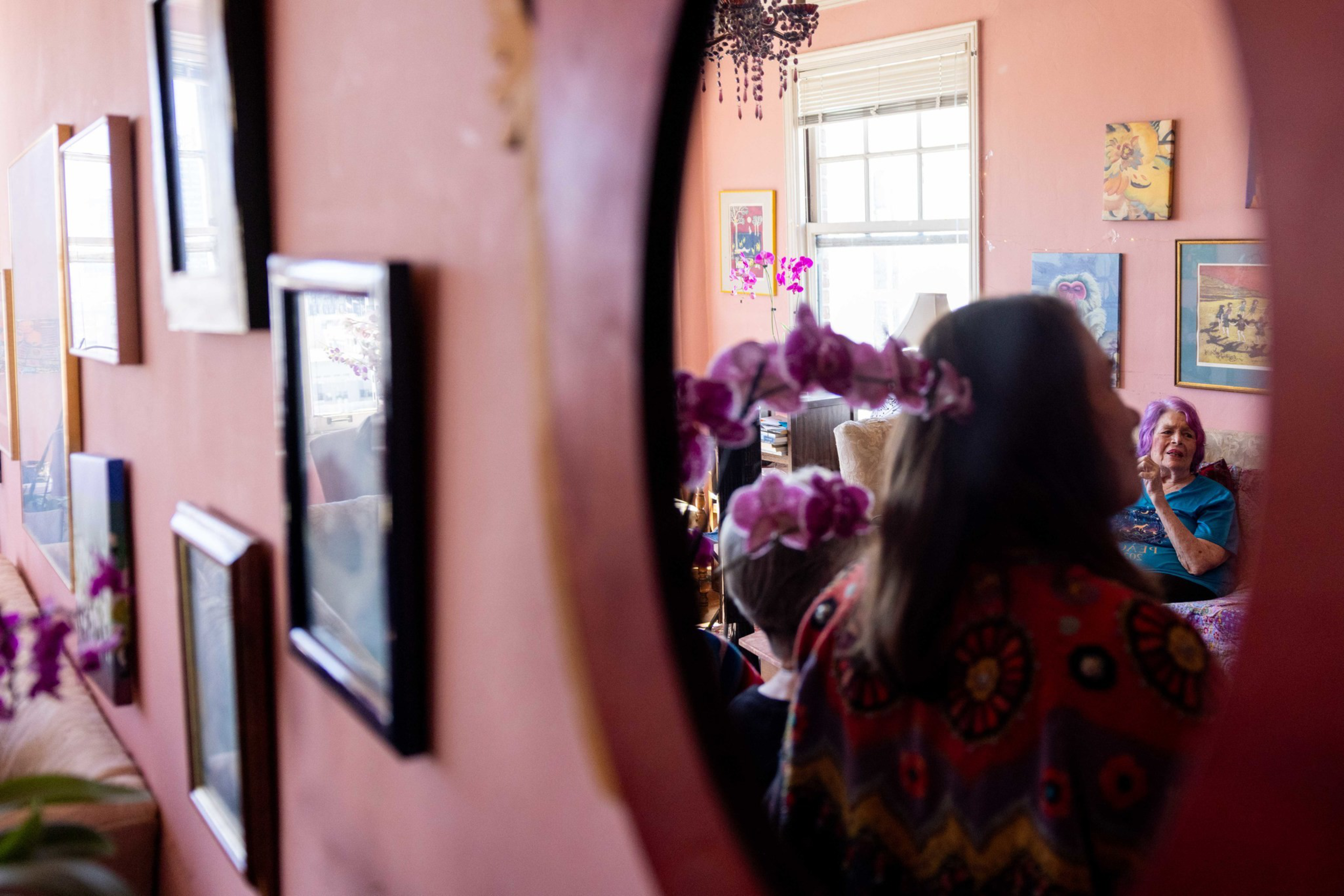 A woman with long hair faces a smiling elderly woman sitting in a cozy room, seen through a round mirror on a pink wall decorated with framed pictures.