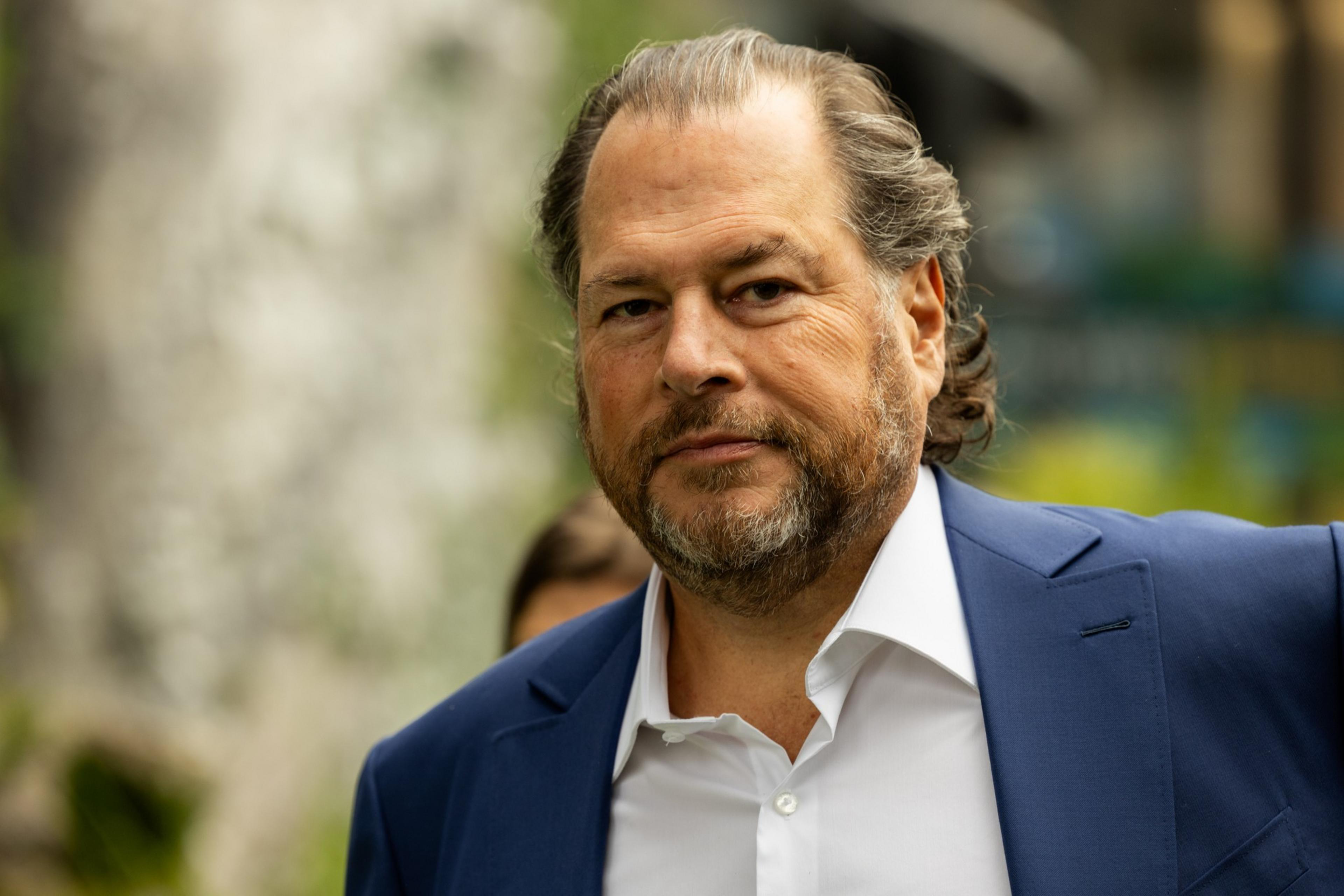 A man with long, dark hair tied back, wearing a blue suit and white shirt, looks seriously at the camera, standing outdoors by greenery.