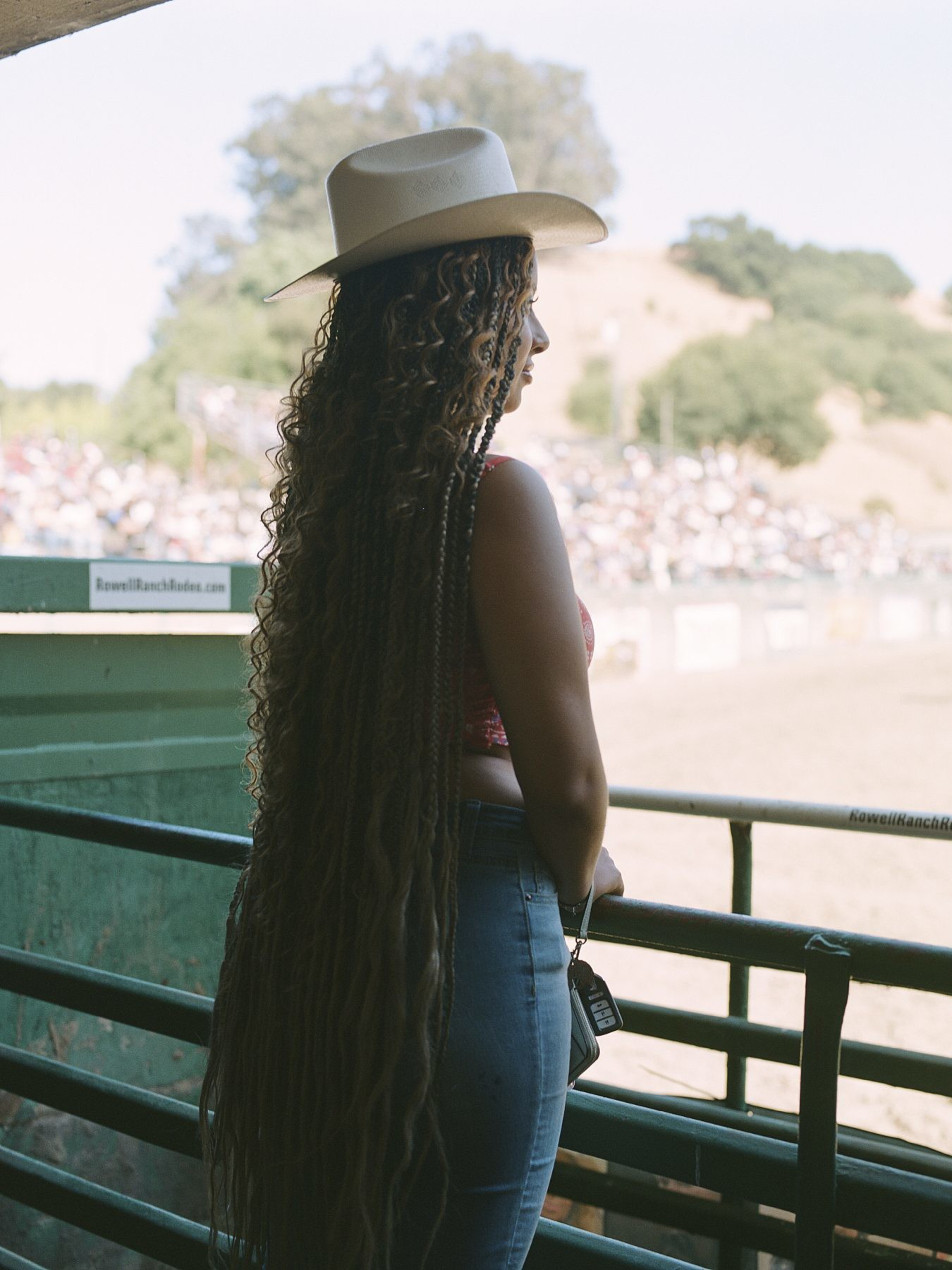 A woman in a hat stands by a railing, facing a large outdoor arena. She has long, curly hair that falls to her waist. The background shows a hillside and spectators.
