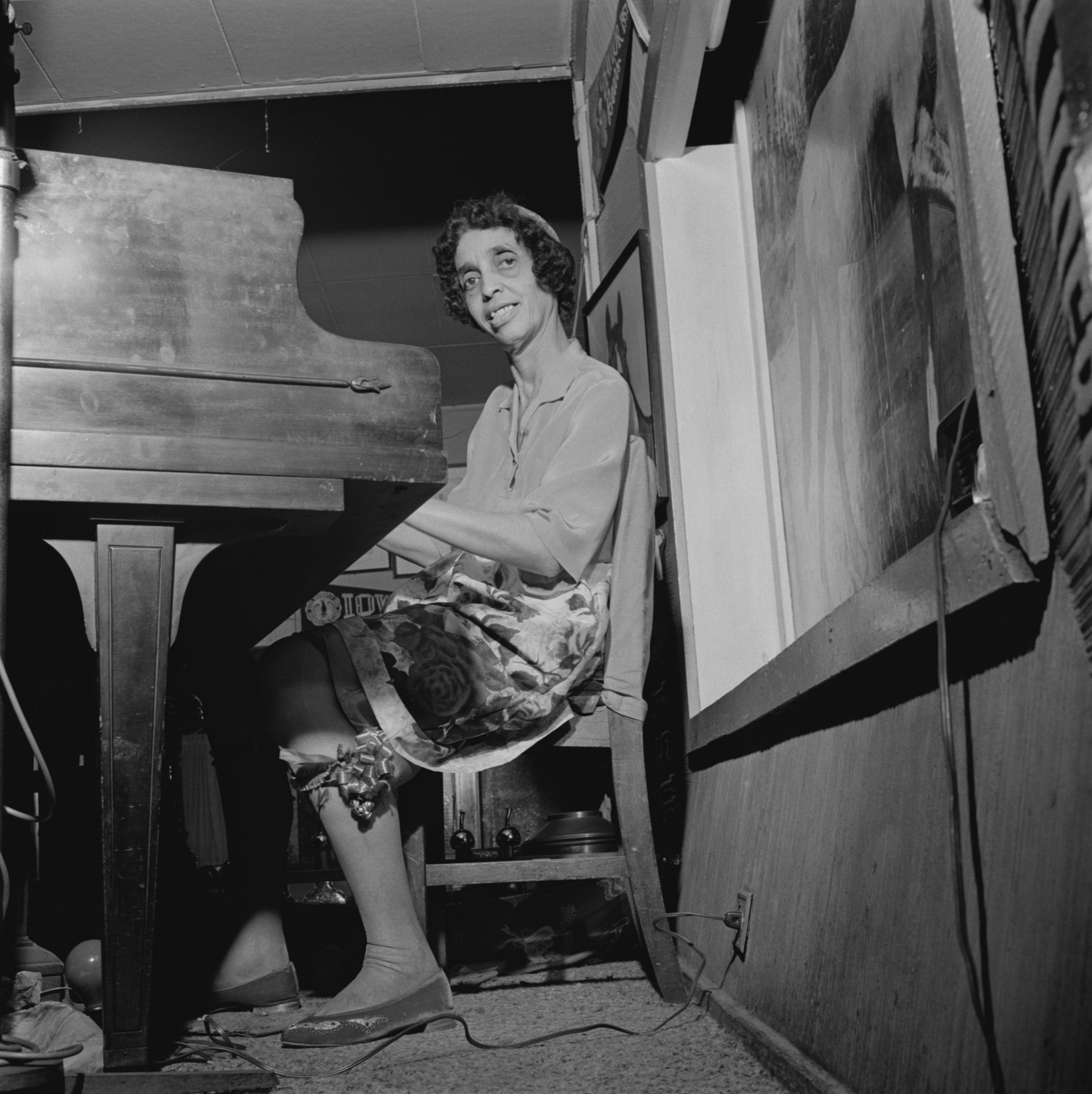 A person sits at a piano, smiling and playing. They wear a floral skirt and blouse in a cozy room with wall art and a carpeted floor.