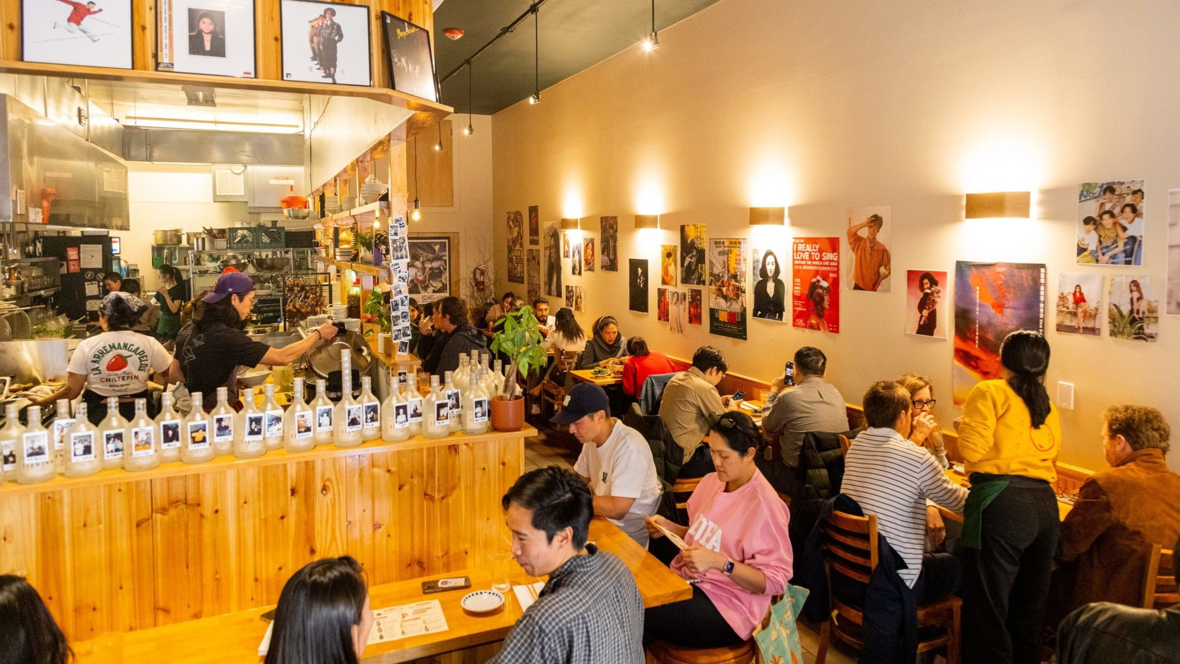 A cozy restaurant with wooden décor is bustling with people dining. Walls are adorned with colorful posters, and the open kitchen is visible in the background.