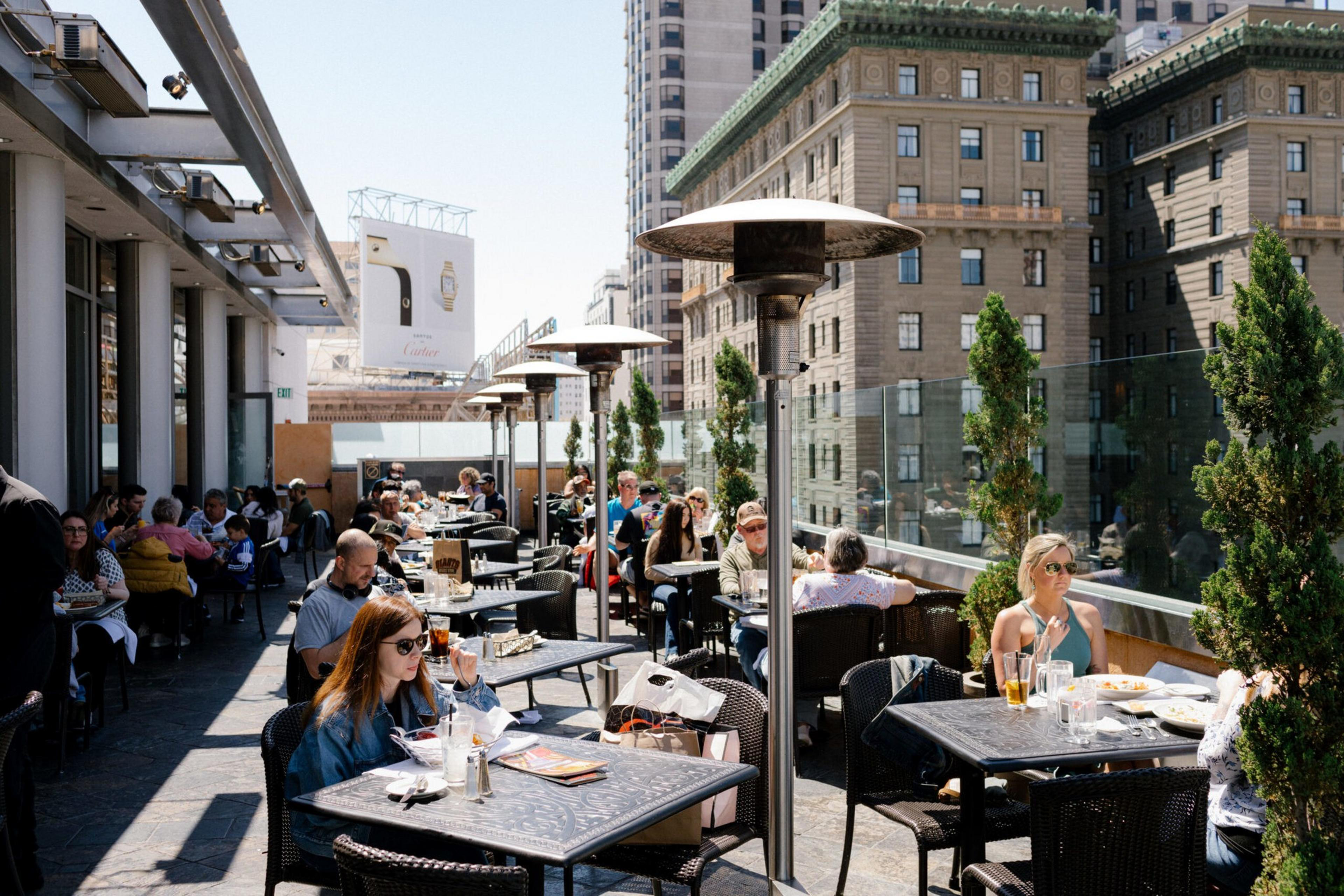 People sit at tables on a sunny outdoor rooftop patio, surrounded by city buildings and tall planters, enjoying food and drinks.