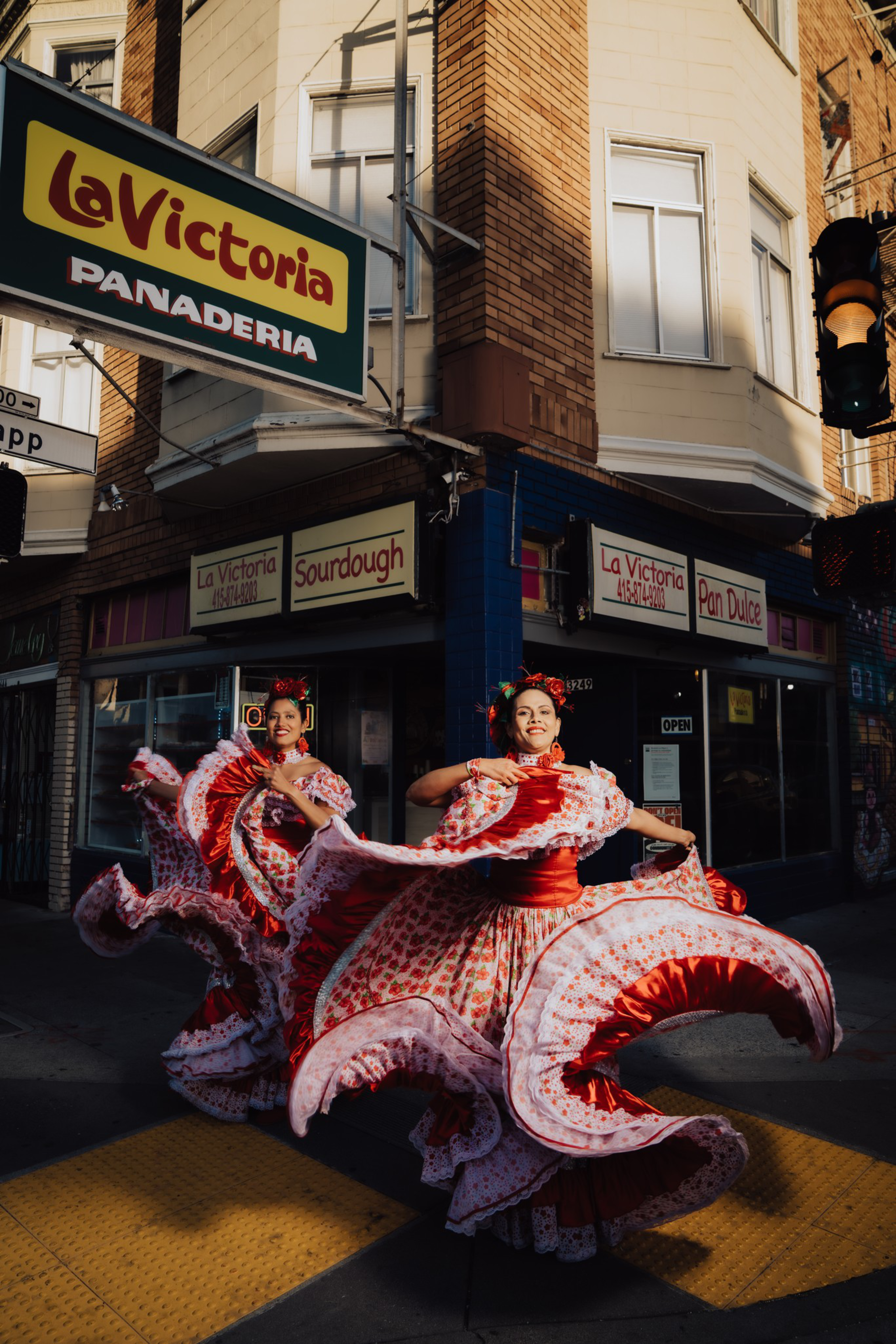 Two women in red and white traditional dresses dance energetically in front of La Victoria Panaderia bakery on a sunny street corner.