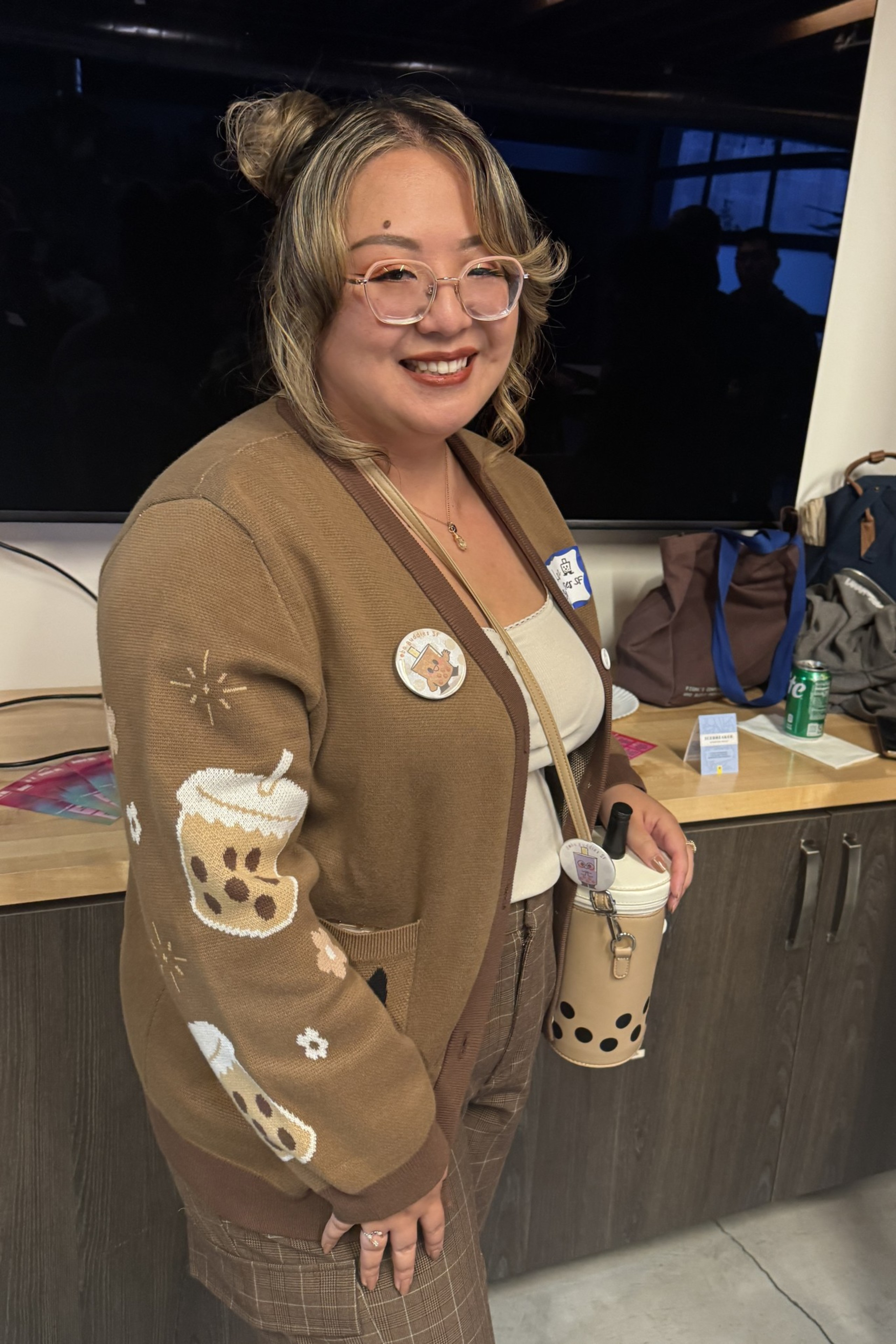A person with glasses and twin buns wears a brown cardigan featuring bubble tea illustrations. They’re holding a matching bubble tea-themed accessory and smiling.