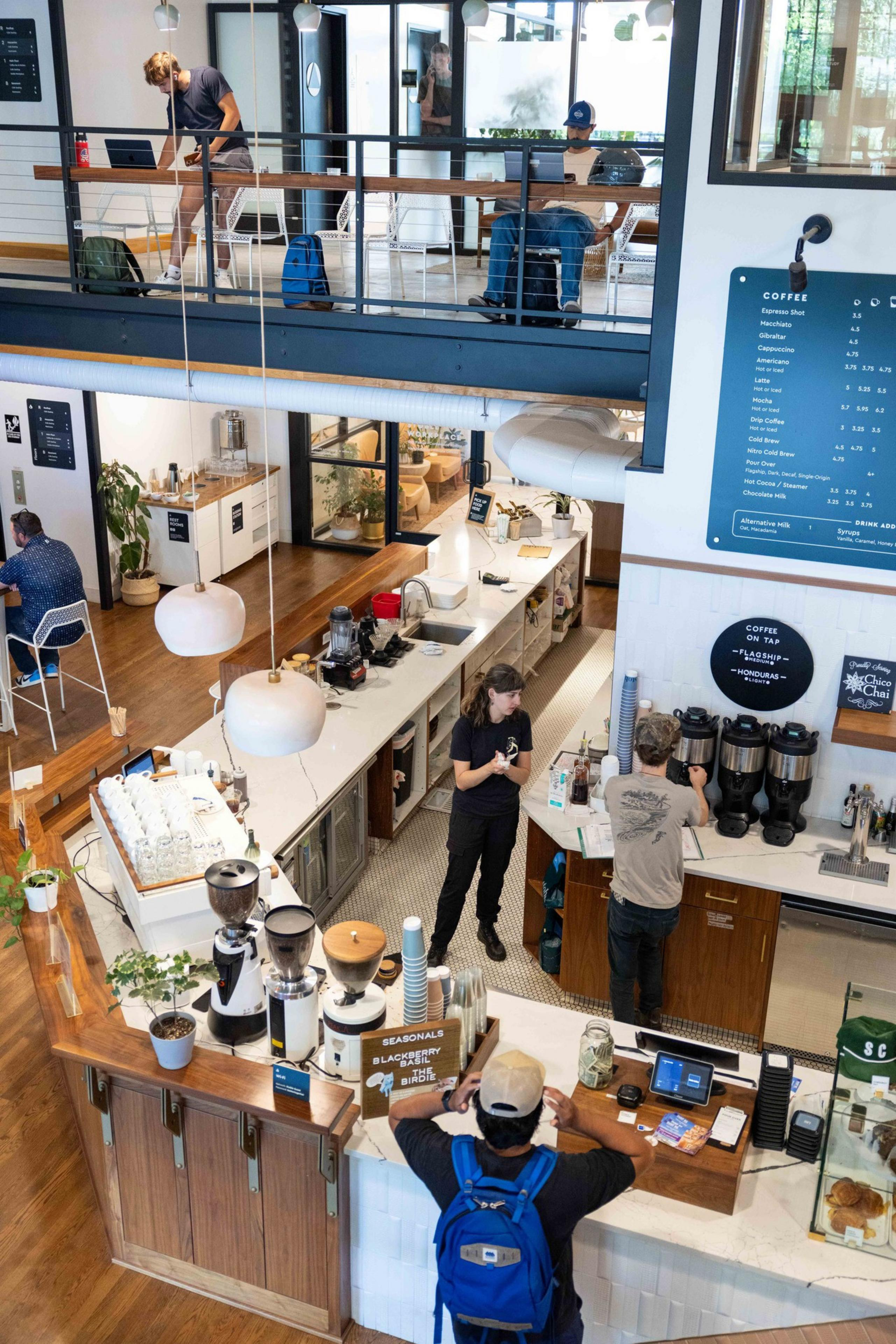 The image shows a modern café with customers at the counter and an upper seating area. Baristas are preparing drinks. A menu is visible on the wall.