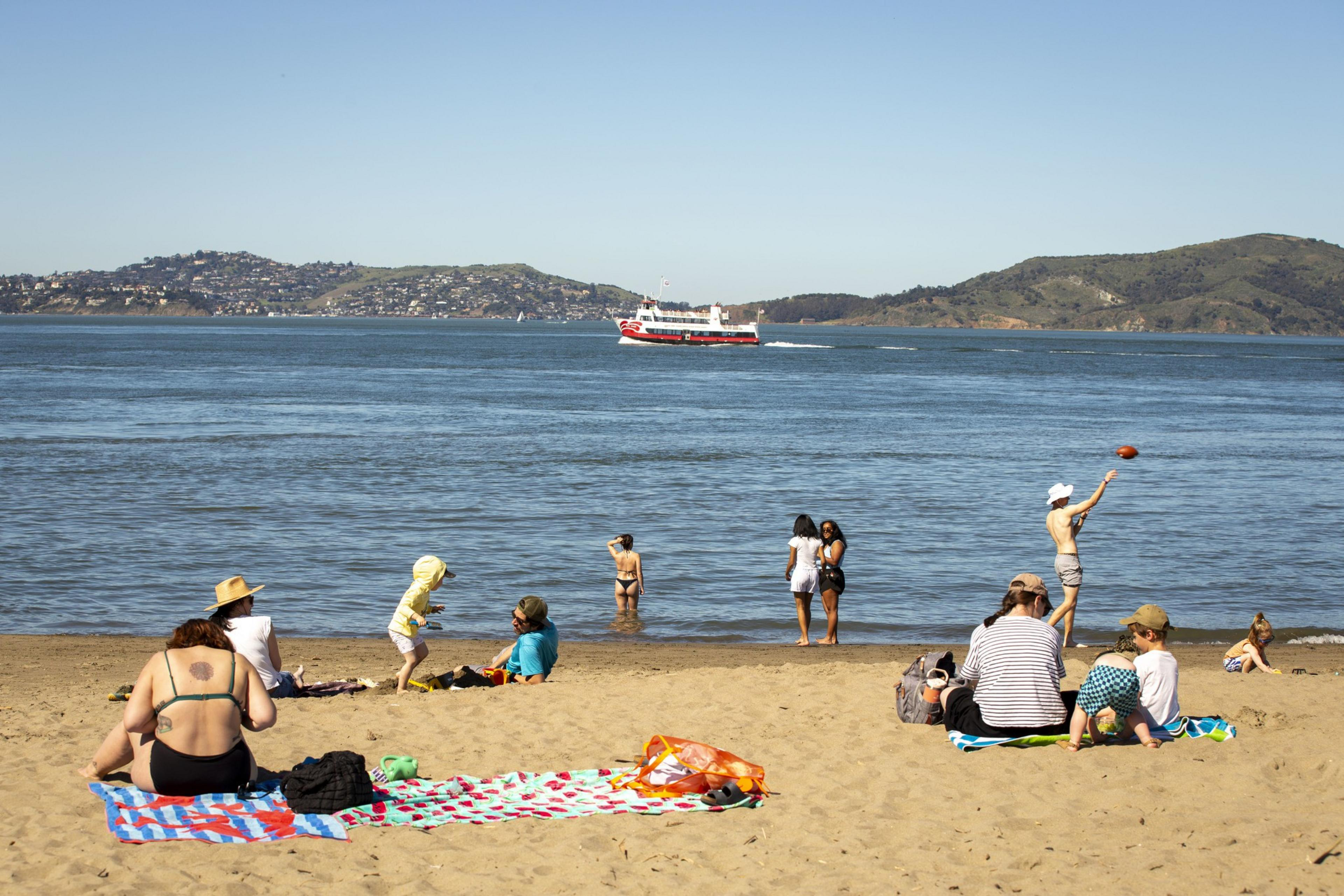 People relax on a sandy beach with towels under clear skies. Some are near the water, while a red and white boat passes by on the sea, with hills in the background.