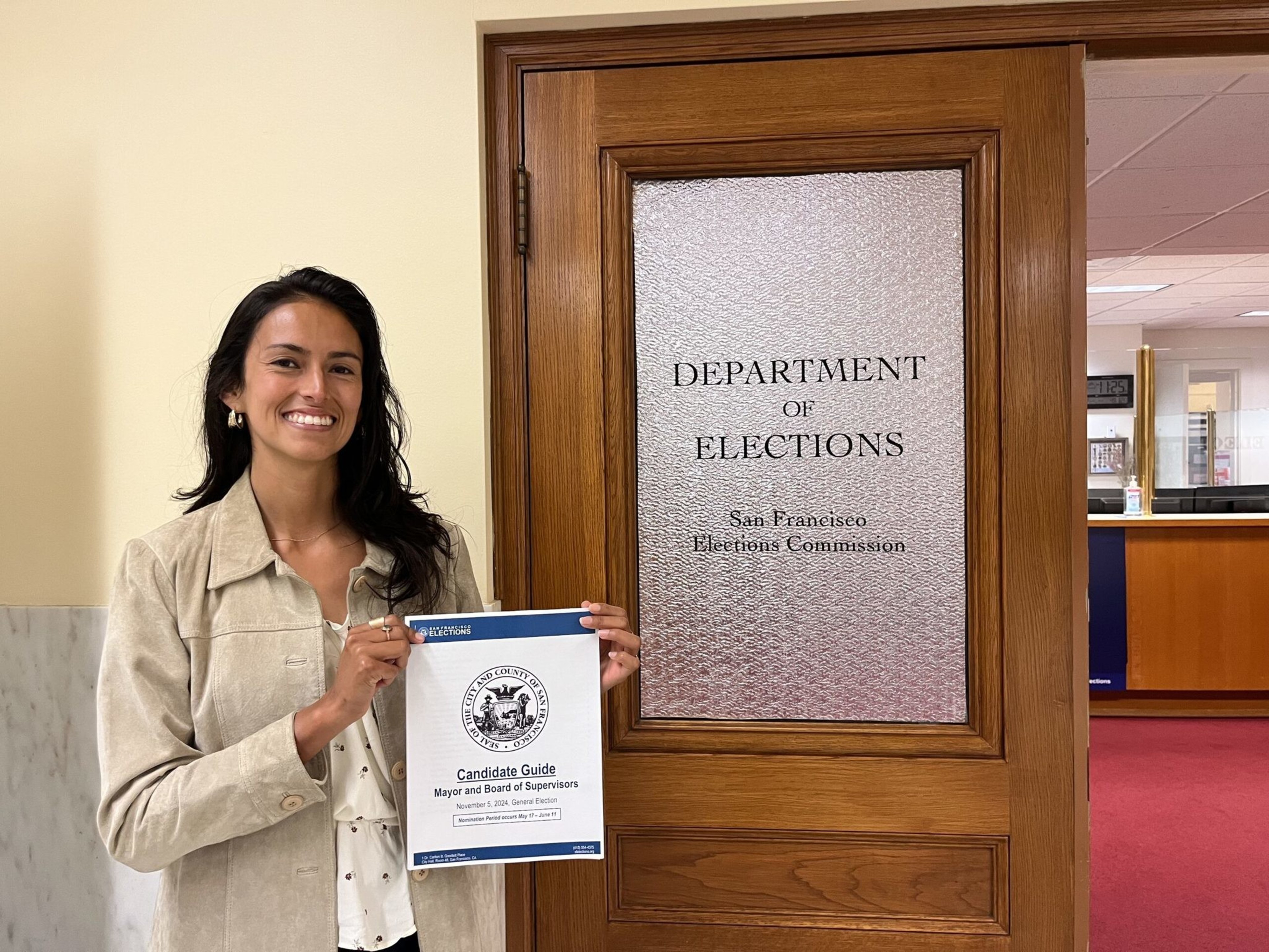 A smiling woman stands next to a wooden door labeled "Department of Elections," holding a candidate guide for San Francisco mayor and board supervisors.