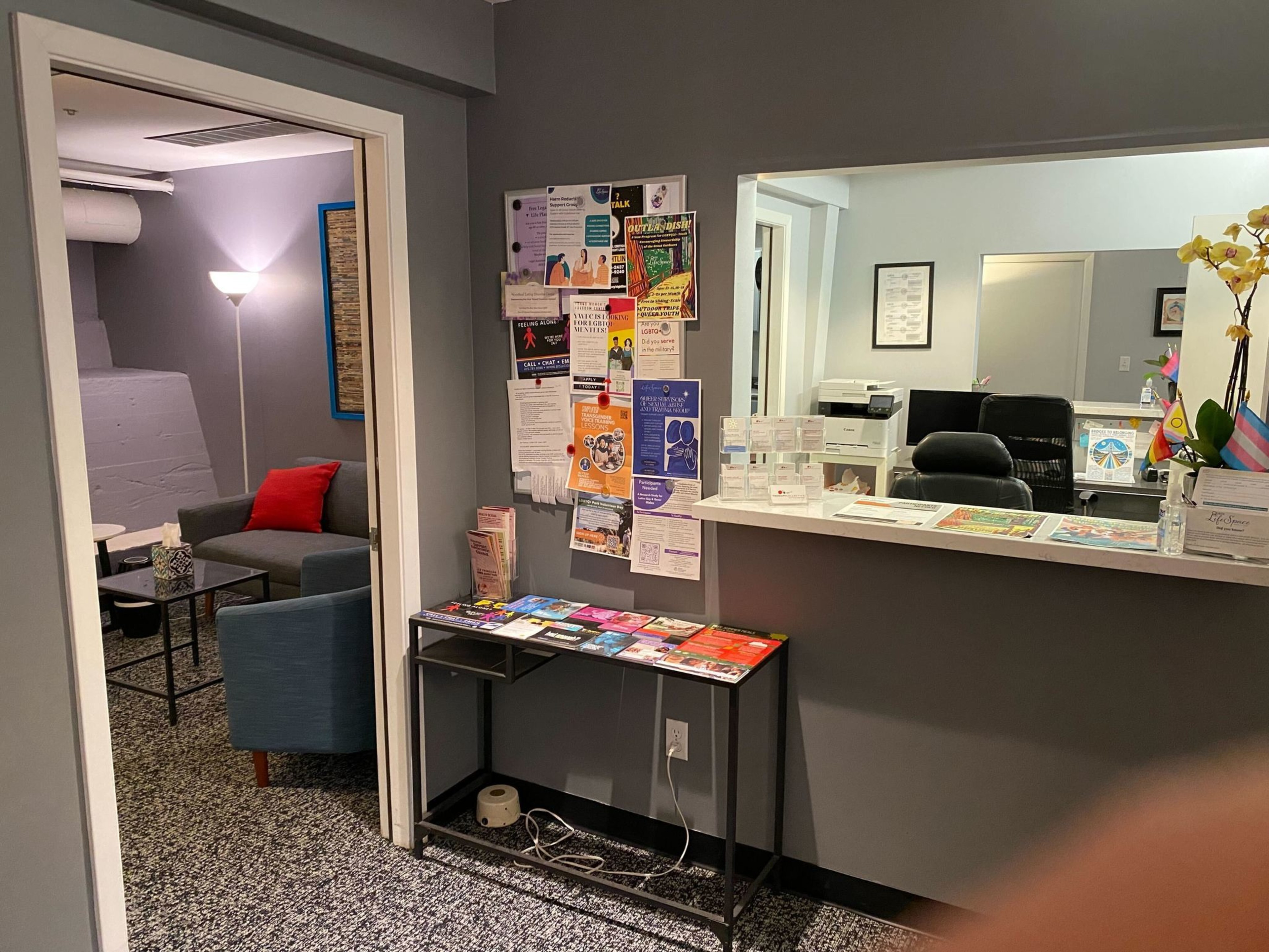 An office lobby with a reception desk, brochures on tables, posters on the wall, and a small seating area with a couch and table lamp.