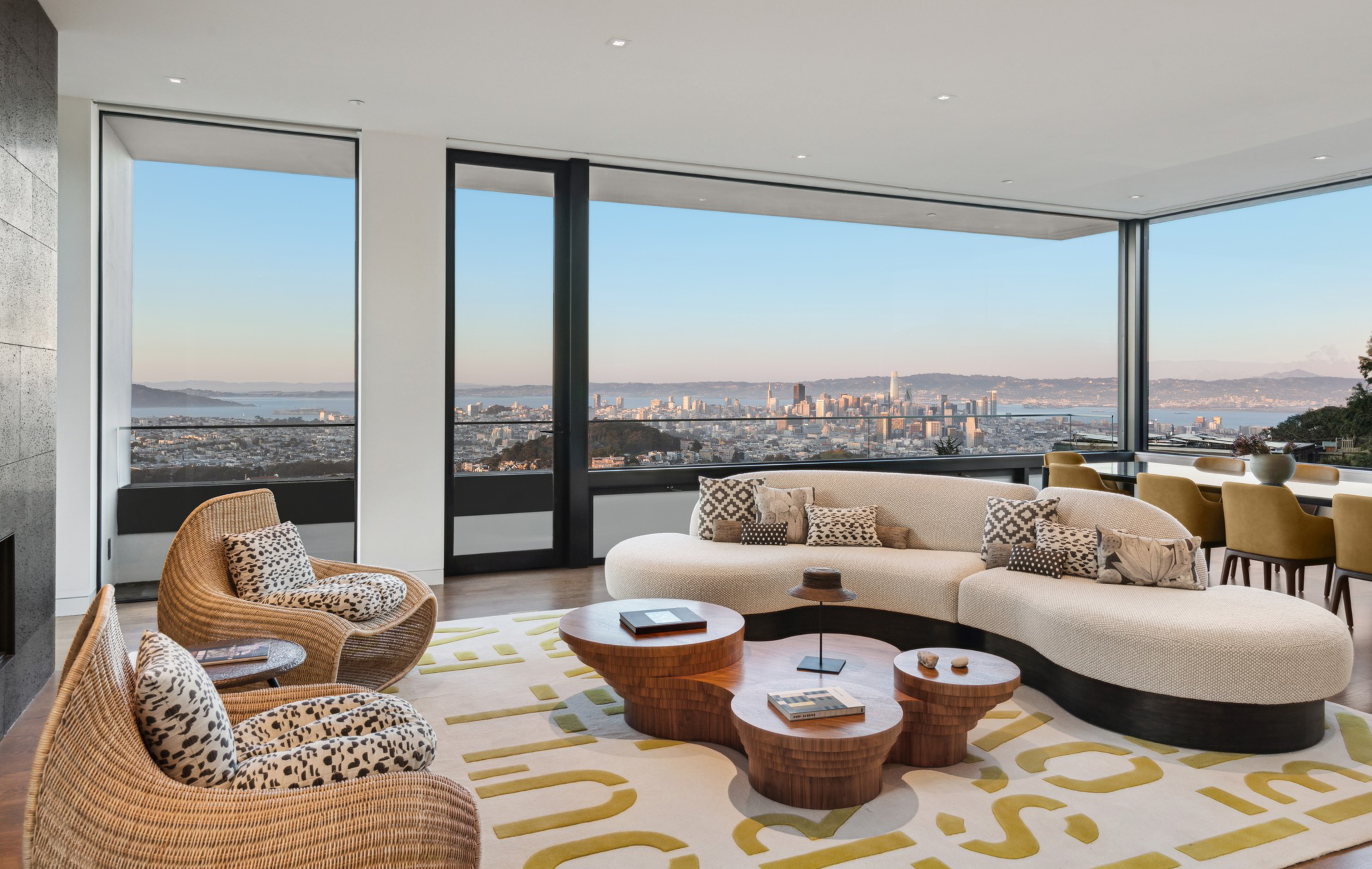 A modern living room with curved beige sofa, patterned cushions, wicker chairs, wooden round tables, and large windows overlooking a city skyline at sunset.