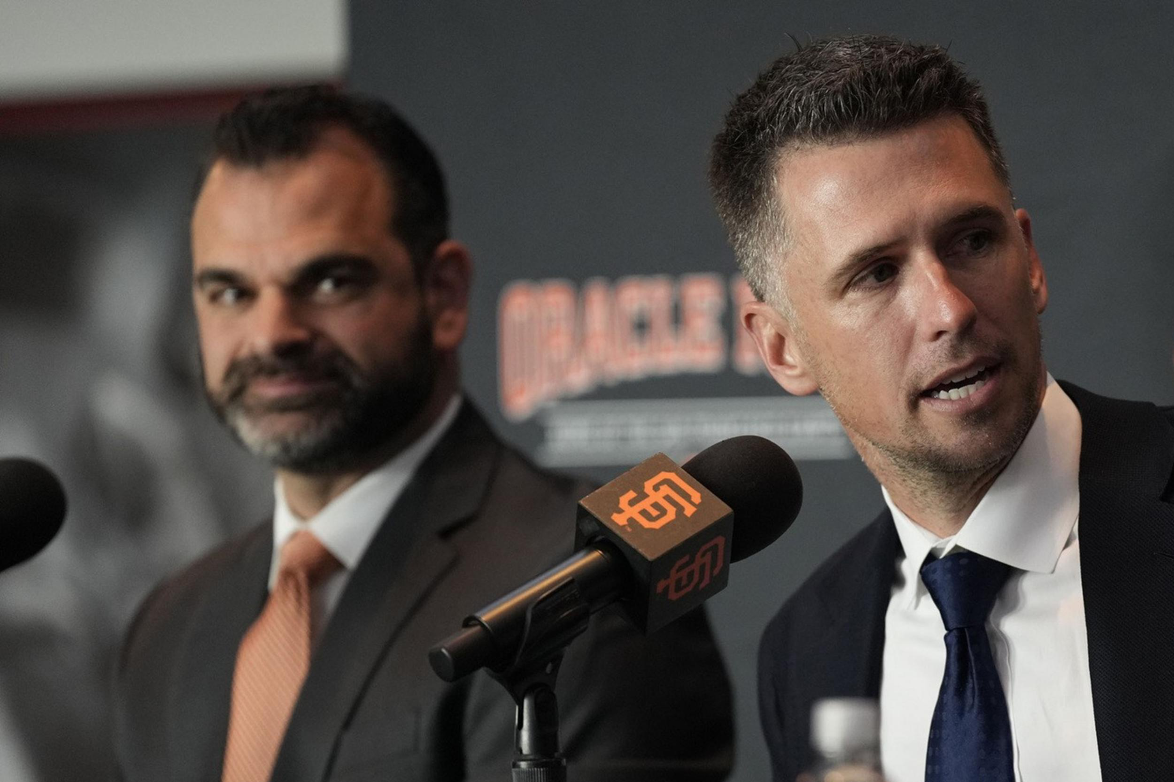 Two men in suits sit at a table with microphones labeled "Giants" in front of them. The man on the right is speaking while gesturing with his hand.