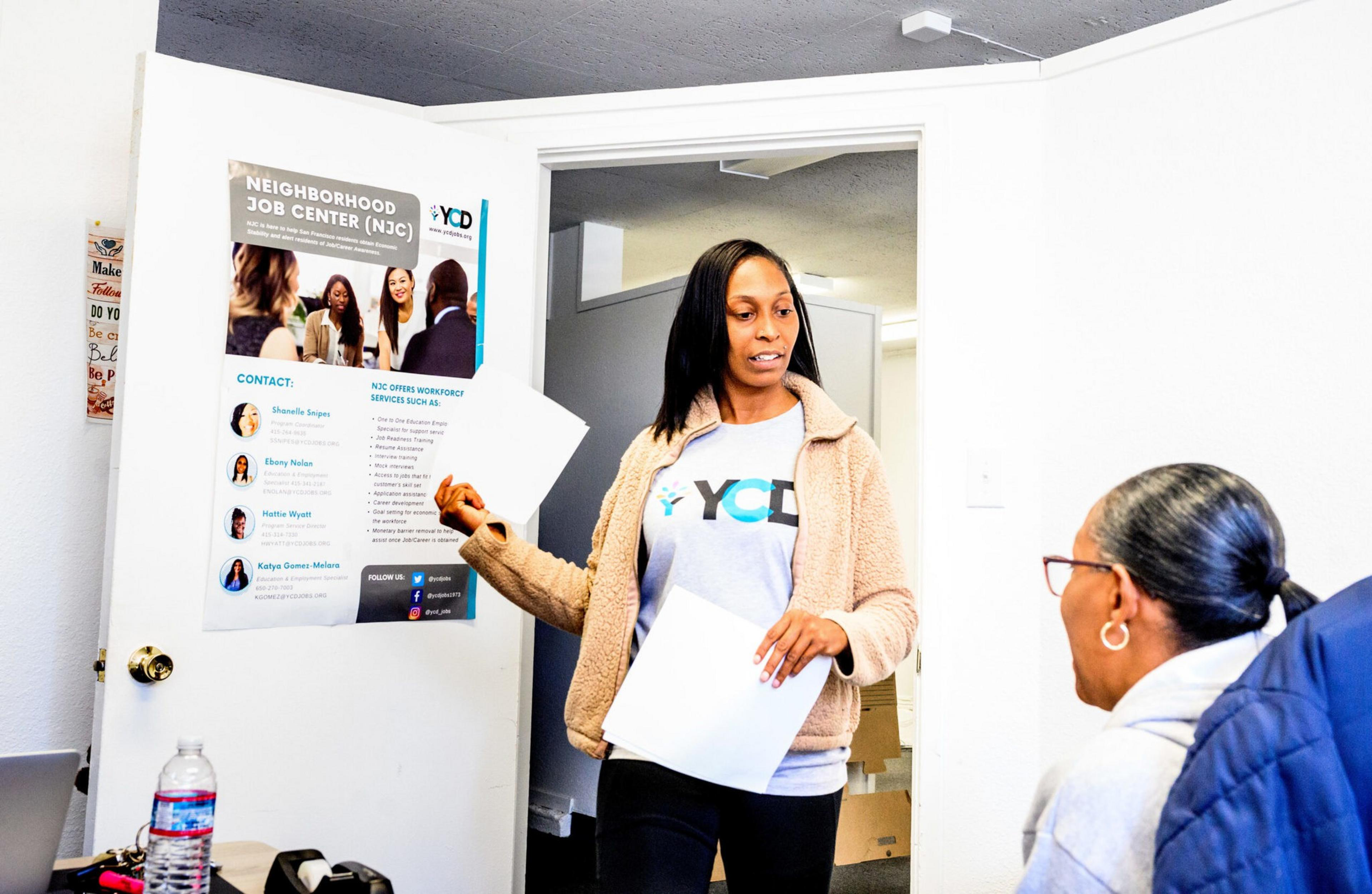 A woman stands holding papers and talking to a seated older woman in an office with a Neighborhood Job Center poster on the wall.