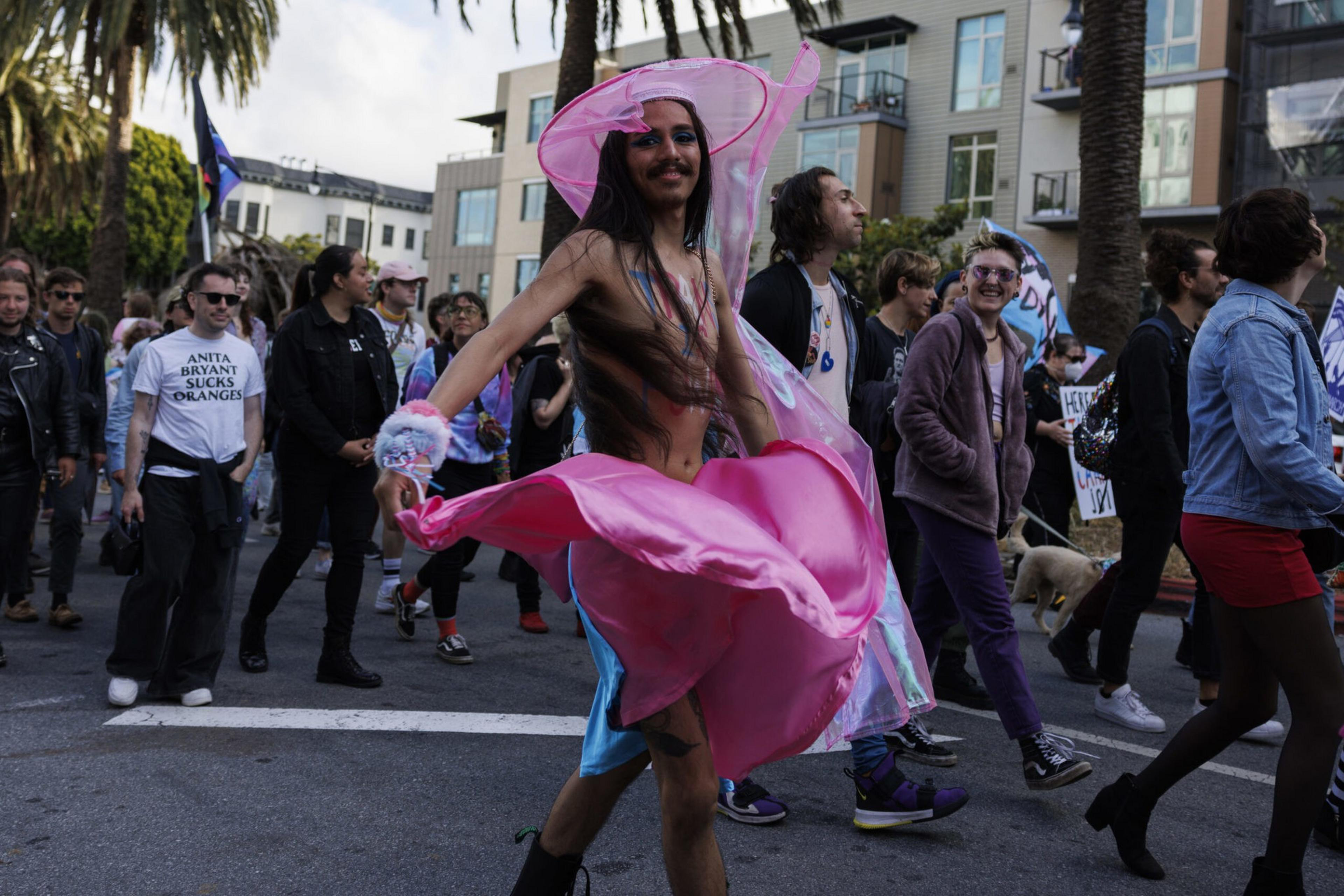 A person wearing a flowing pink and blue outfit twirls in the street amidst a diverse crowd of people walking along a city sidewalk.