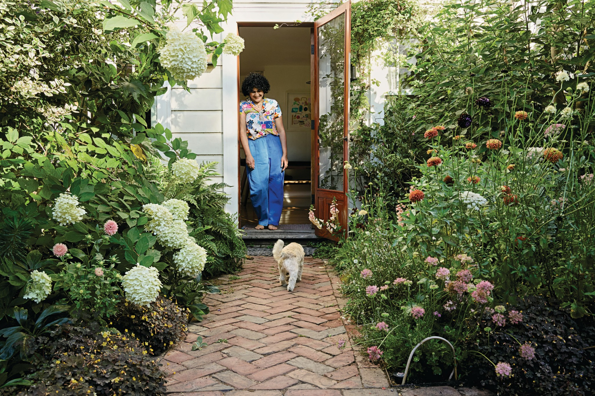 A woman in a floral shirt and blue pants stands barefoot in an open doorway, watching a small dog walk on a brick path surrounded by lush garden plants.