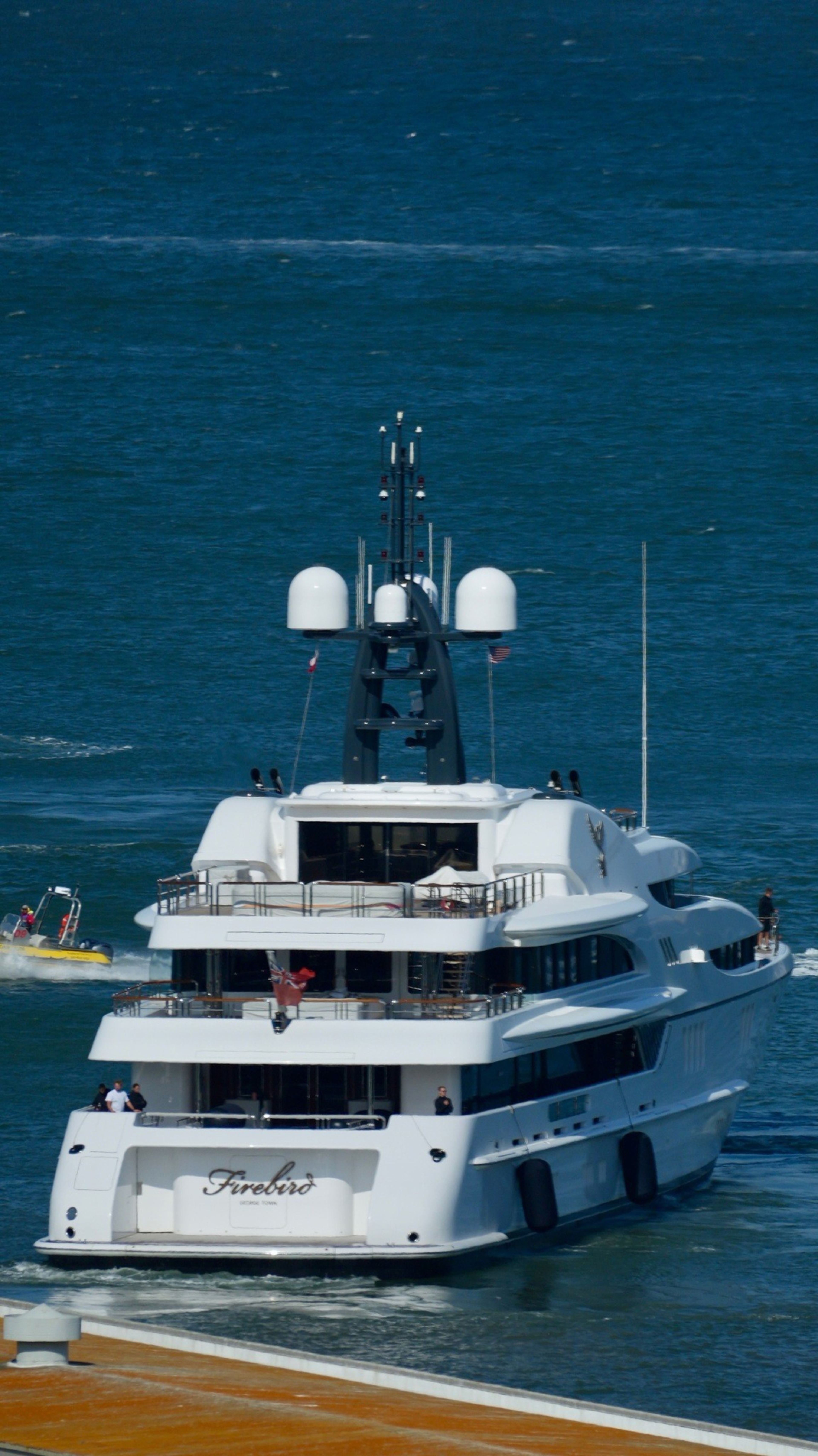 A large white yacht named Firebird sails on blue water, with multiple decks, antennas, and people visible on board.