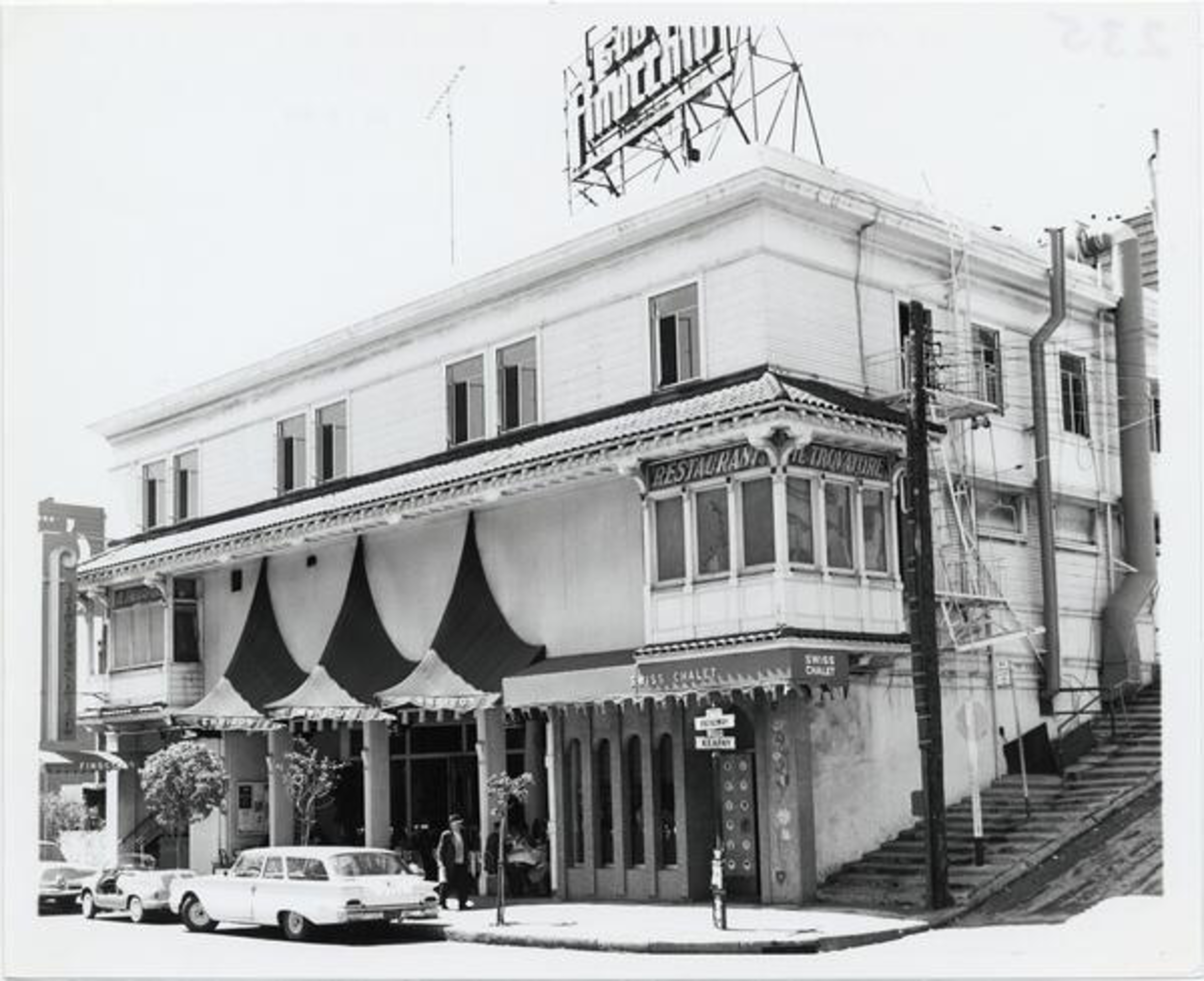 A two-story building with a striped awning houses a restaurant named "Le Tranquille," vintage cars are parked outside on a sloped street.